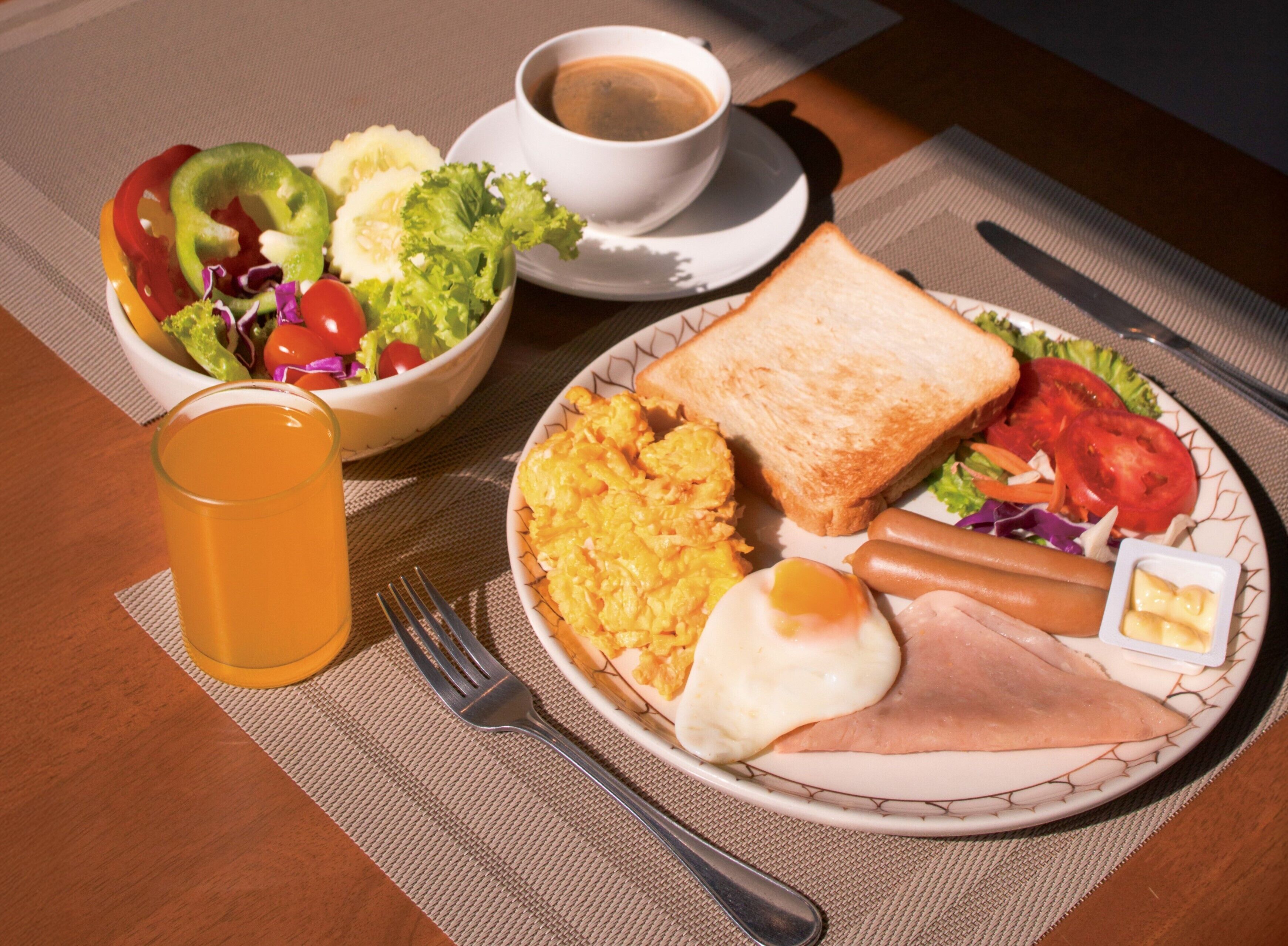 Un plato de desayuno con tostadas, huevos, jamón y una ensalada, una taza de café y un vaso de jugo de naranja sobre una mesa.