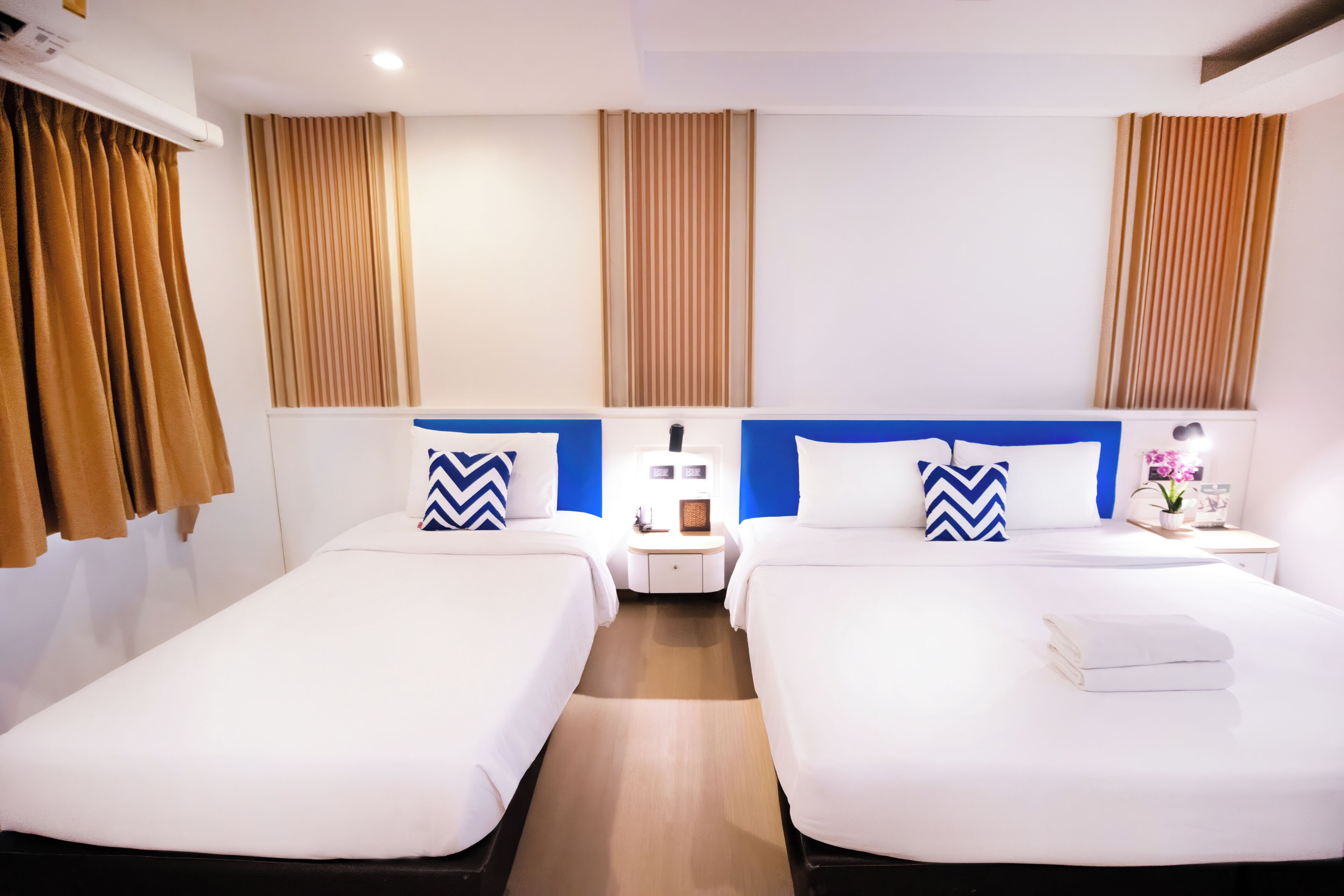 A hotel room with two beds, a nightstand, a lamp, and a window with curtains.