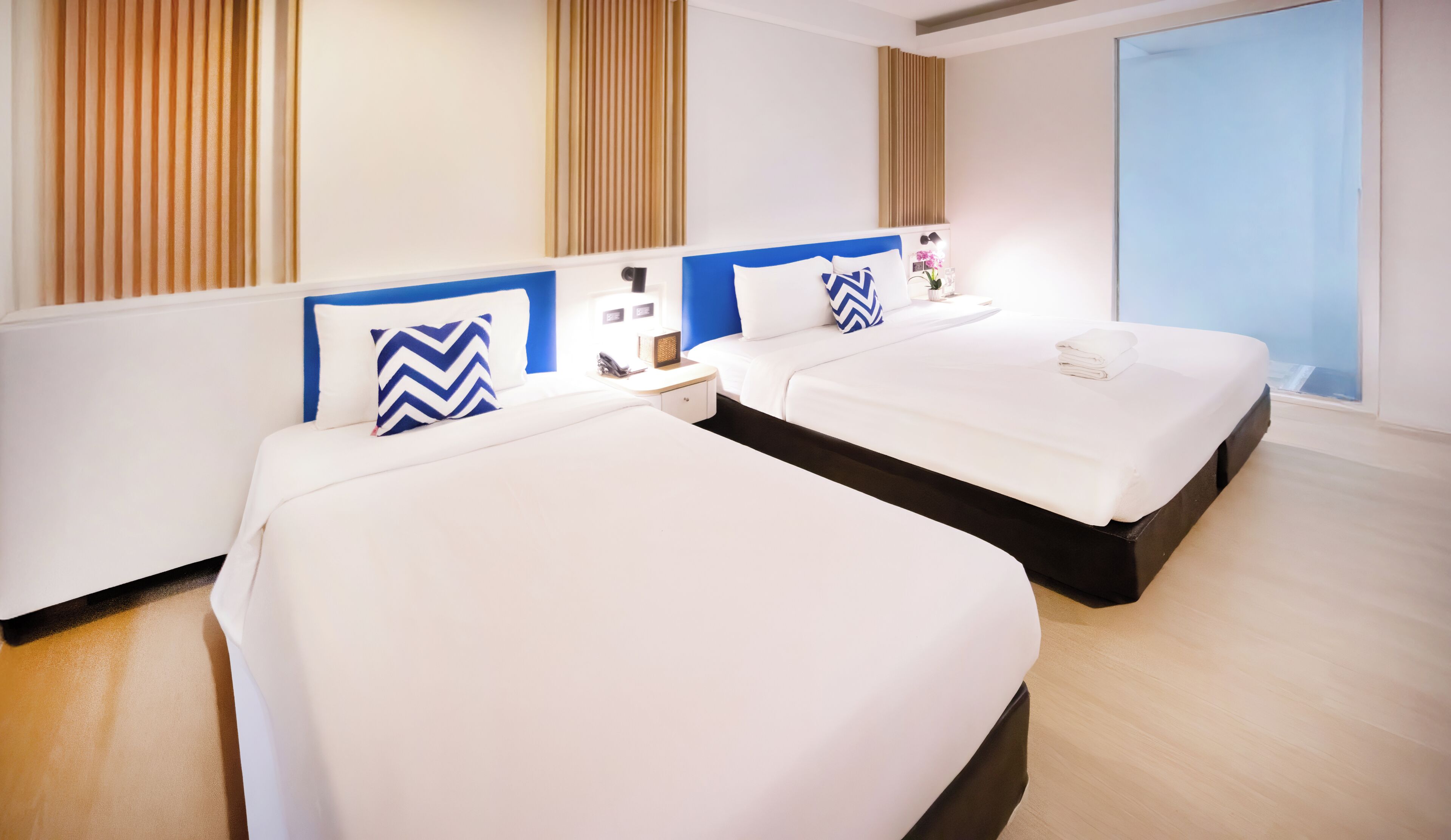 A hotel room with two beds, a bedside table with a lamp, a small table with a vase, and a window with a view.