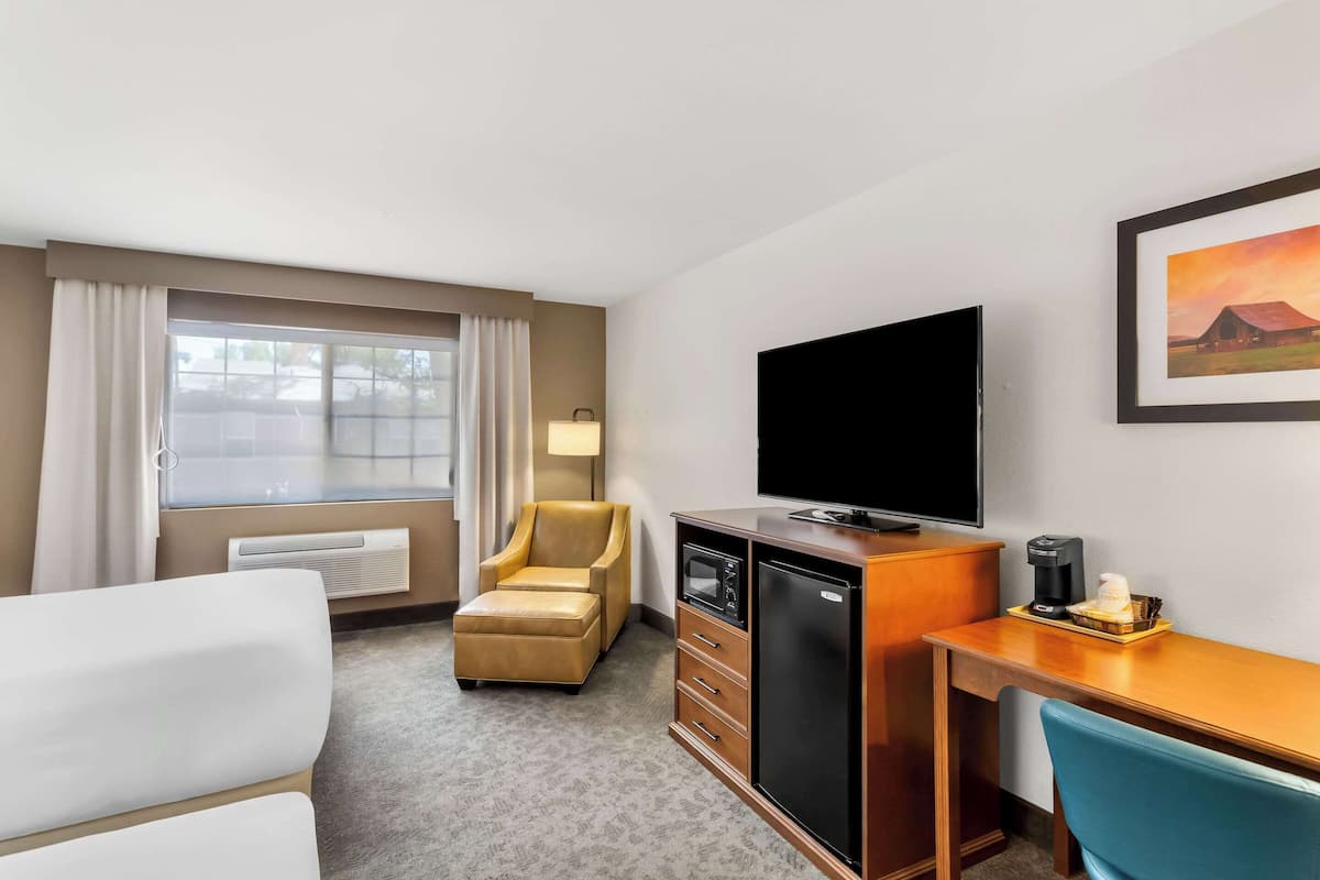 A hotel room with a bed, a desk with a computer, a television, a chair, a small refrigerator, and a window with curtains.
