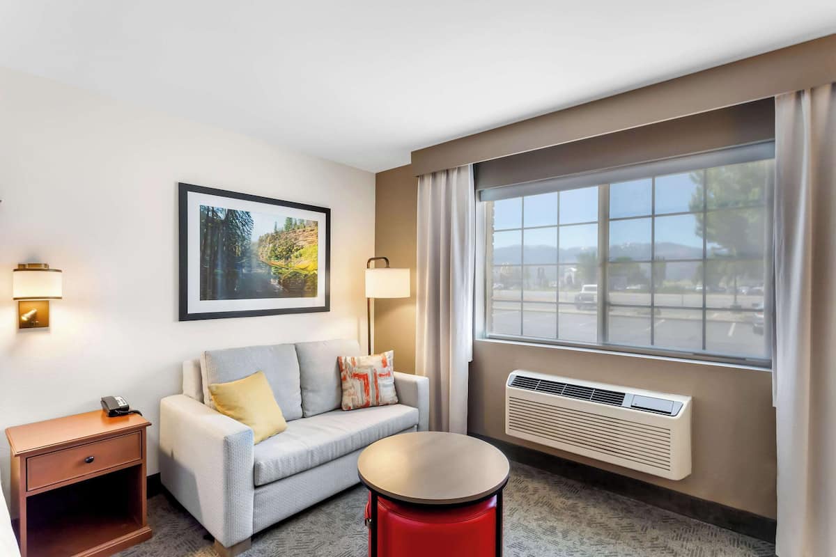 A hotel room with a sofa, a small table, a nightstand, a framed picture on the wall, and a window with a view of the outdoors.
