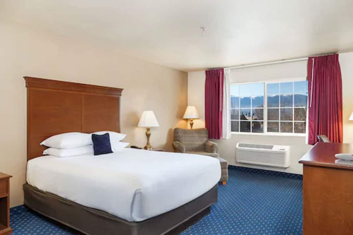 A hotel room with a large bed, a chair, a desk, a window with curtains, and a view of mountains.
