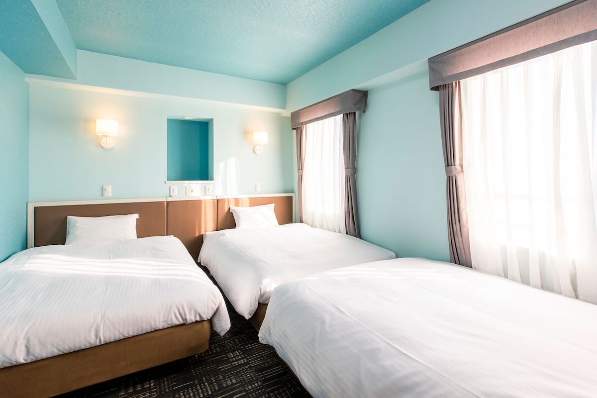 A hotel room with two beds, a large window with curtains, a wall-mounted mirror, and two wall sconces.