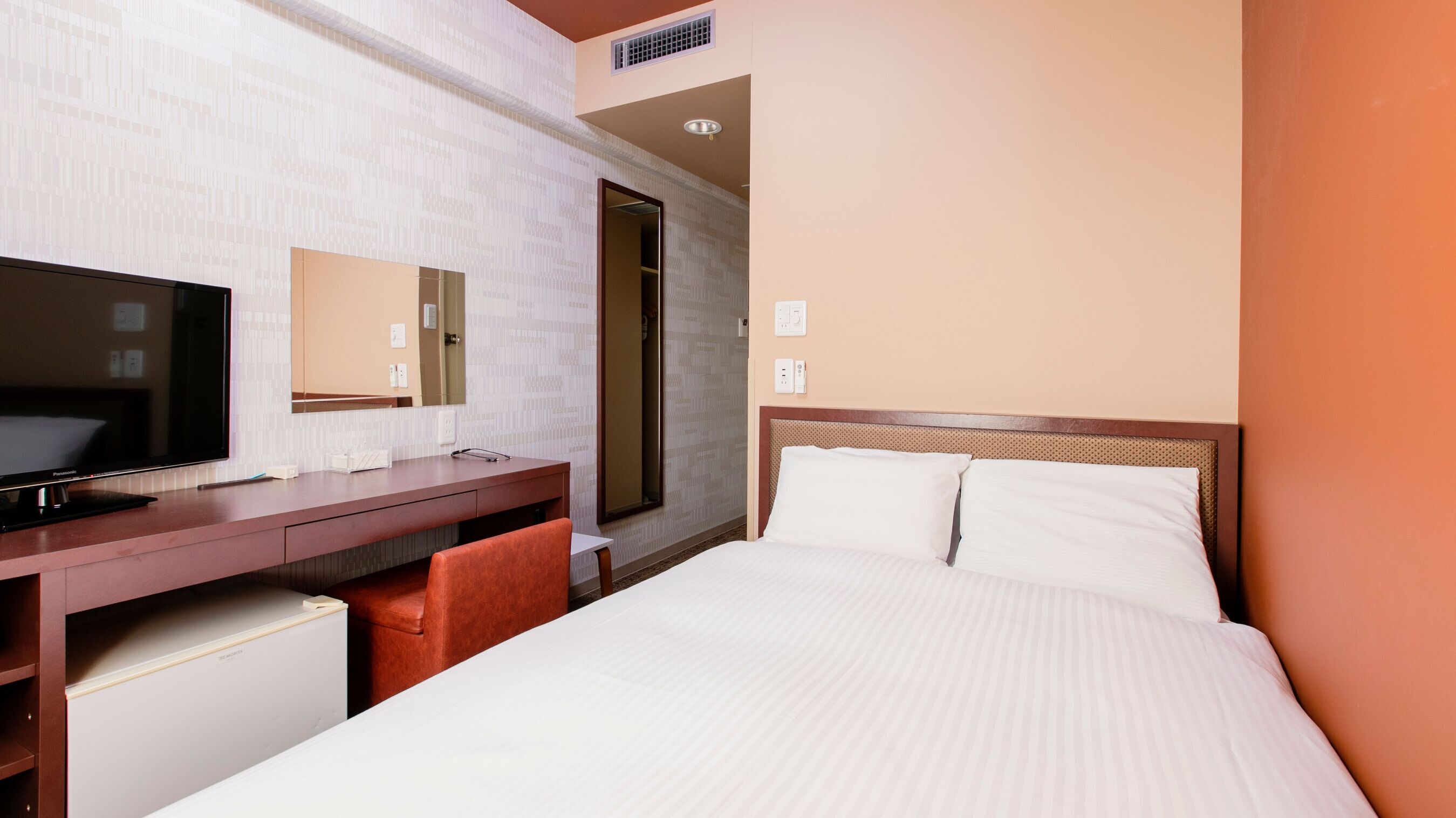A hotel room with a bed, a desk with a flat-screen TV, a mirror, and a door leading to a bathroom.