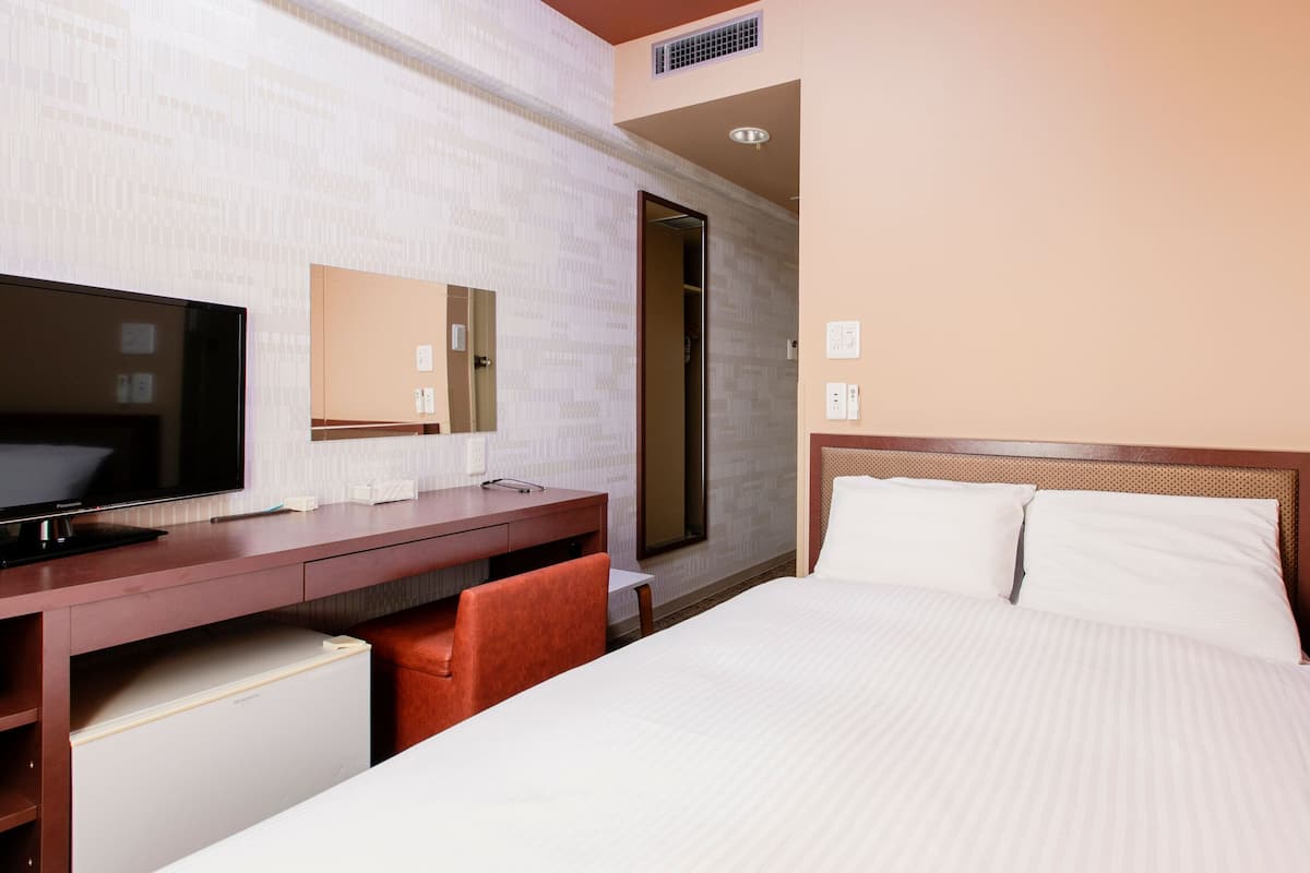 A hotel room with a bed, a desk with a flat-screen TV, a mirror, and a door leading to a bathroom.