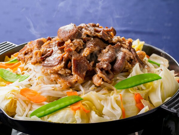 A close-up of a dish with sliced meat, vegetables, and noodles in a black pan.