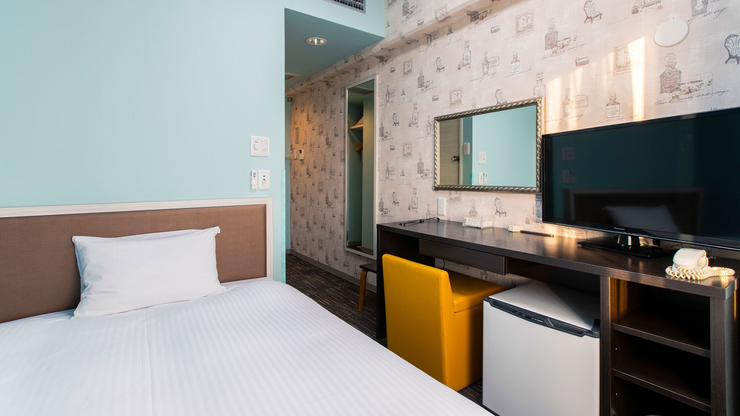 A hotel room with a bed, a desk with a mirror, a small refrigerator, and a yellow chair.