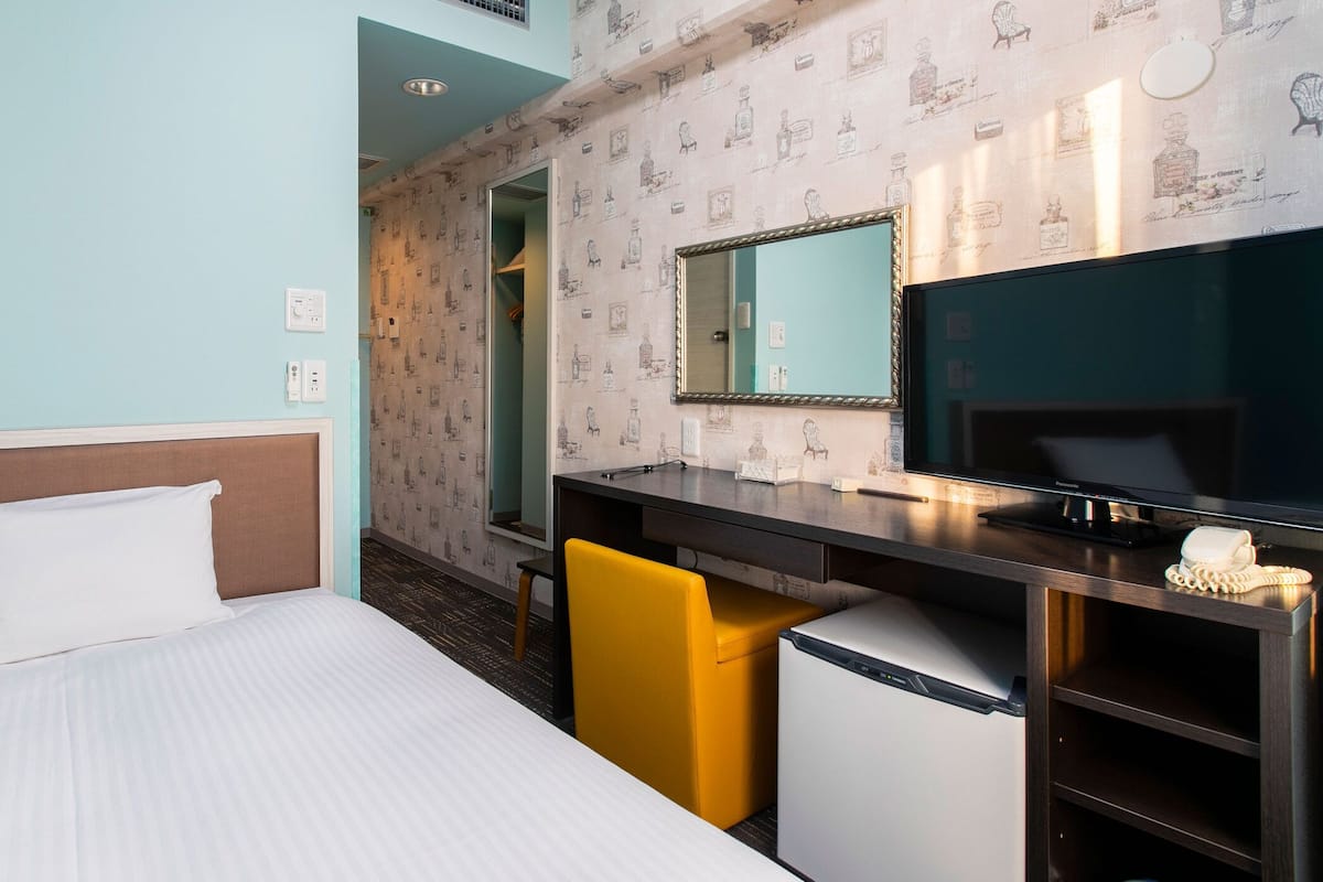 A hotel room with a bed, a desk with a mirror, a small refrigerator, and a yellow chair.