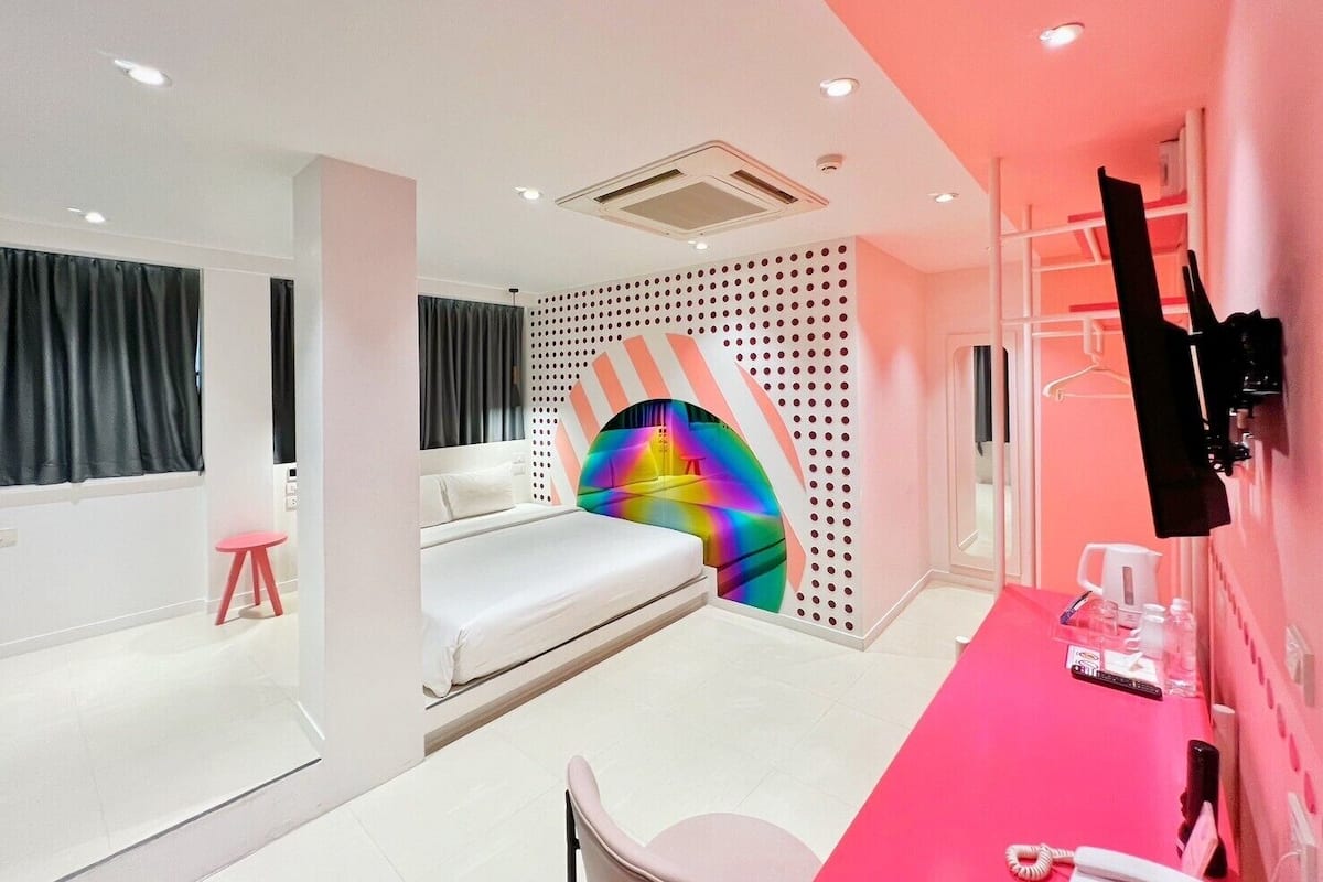 A modern hotel room with a bed, a desk with a pink tablecloth, a TV mounted on the wall, and a colorful wall decoration.