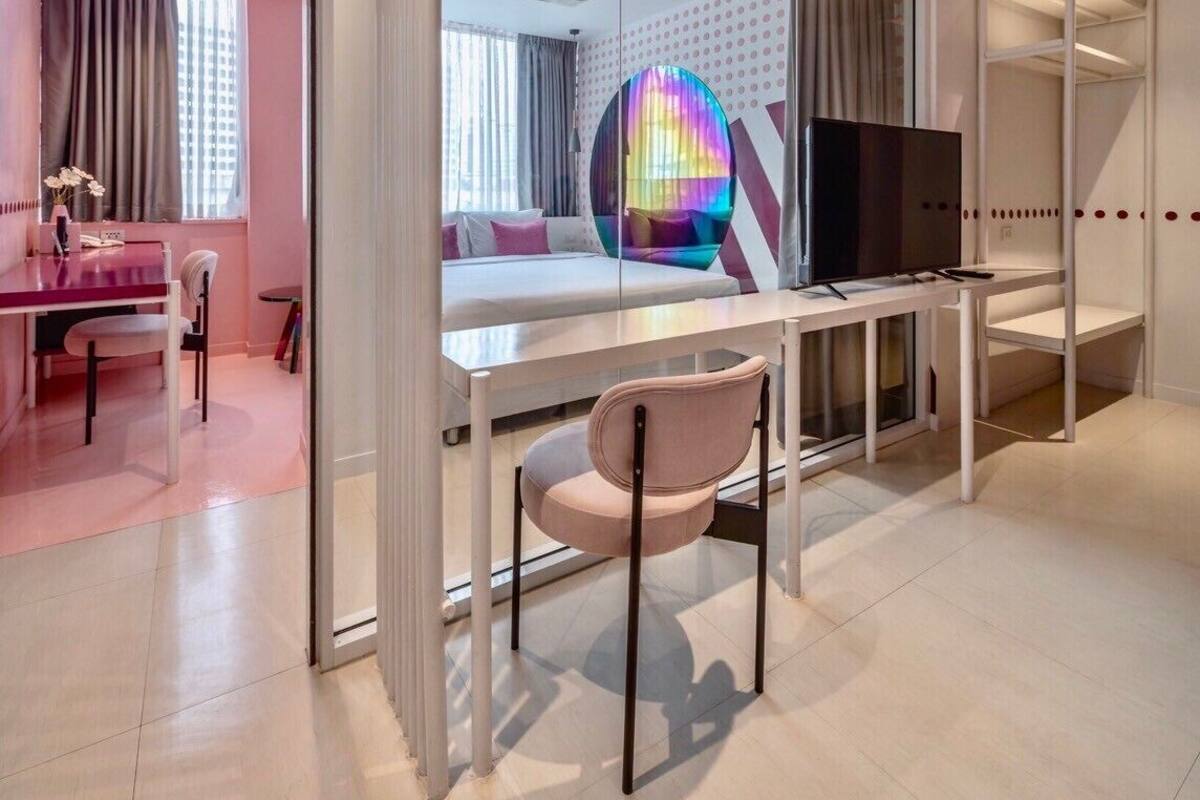 A modern hotel room with a glass partition, a bed, a desk with a chair, a TV, and a wardrobe.