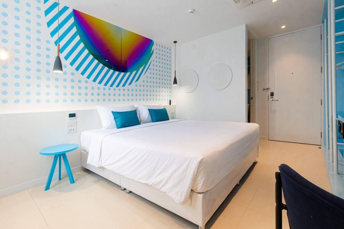 A modern bedroom with a large bed, a small blue table, and a colorful abstract wall art piece.