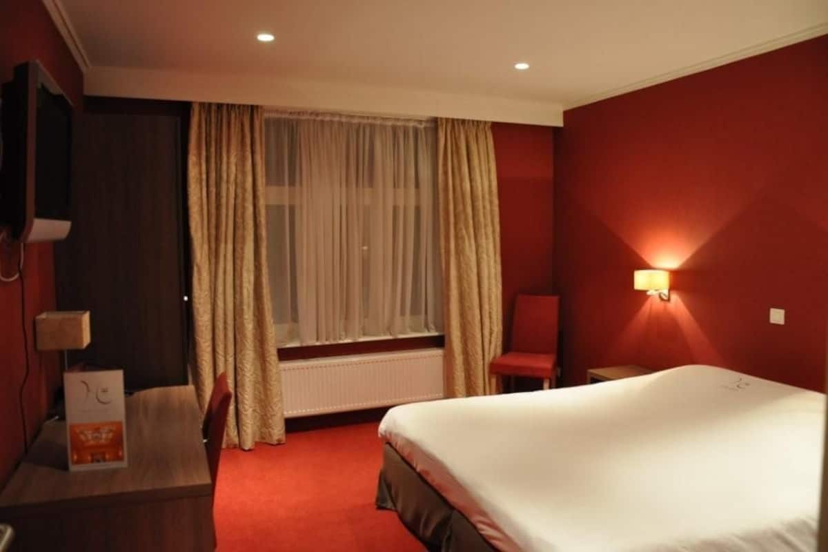 Habitación de hotel con cama, escritorio, silla, televisor y ventana con cortinas.