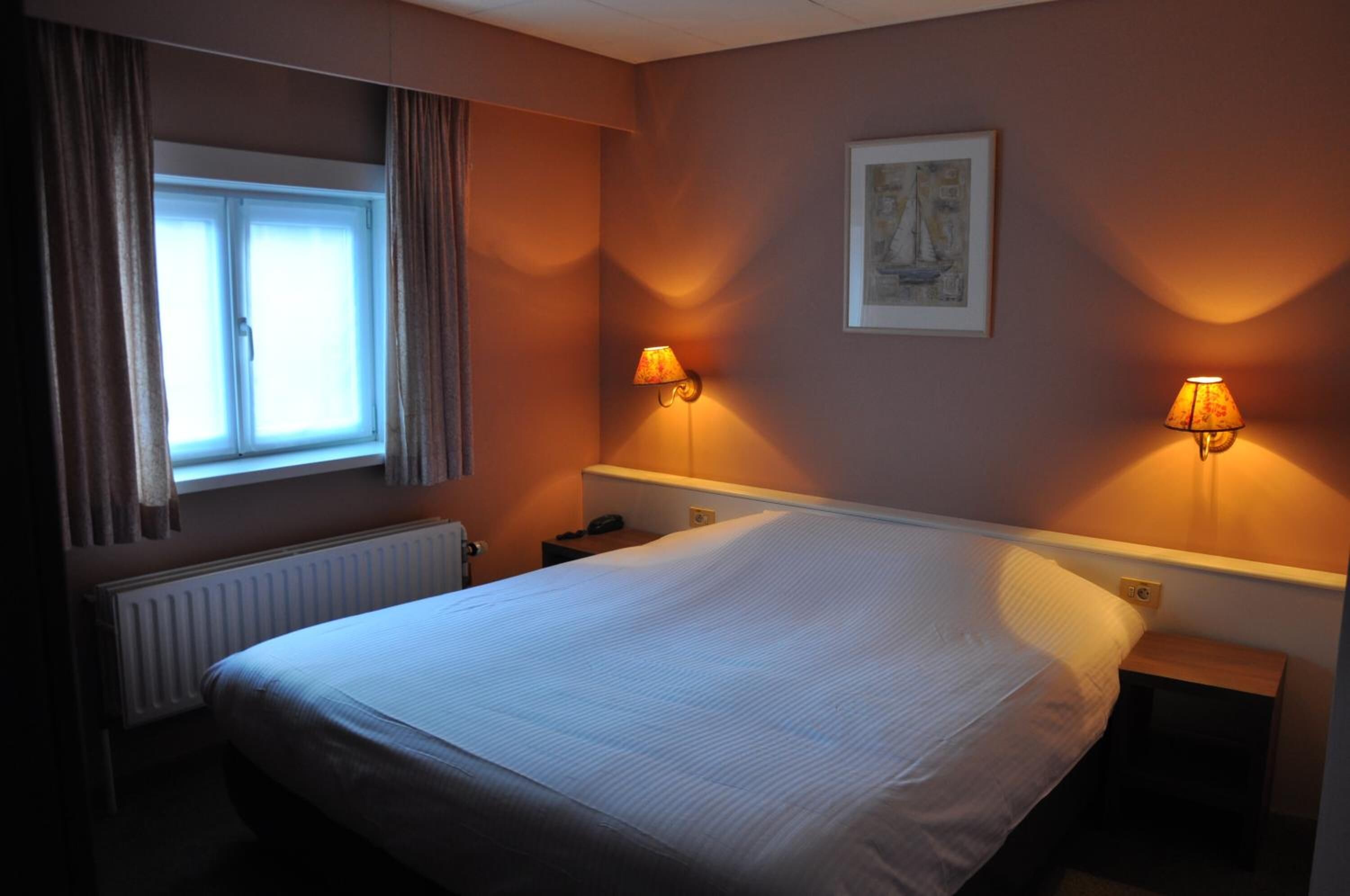 A hotel room with a large bed, two bedside lamps, a window with curtains, a radiator, and a framed picture on the wall.