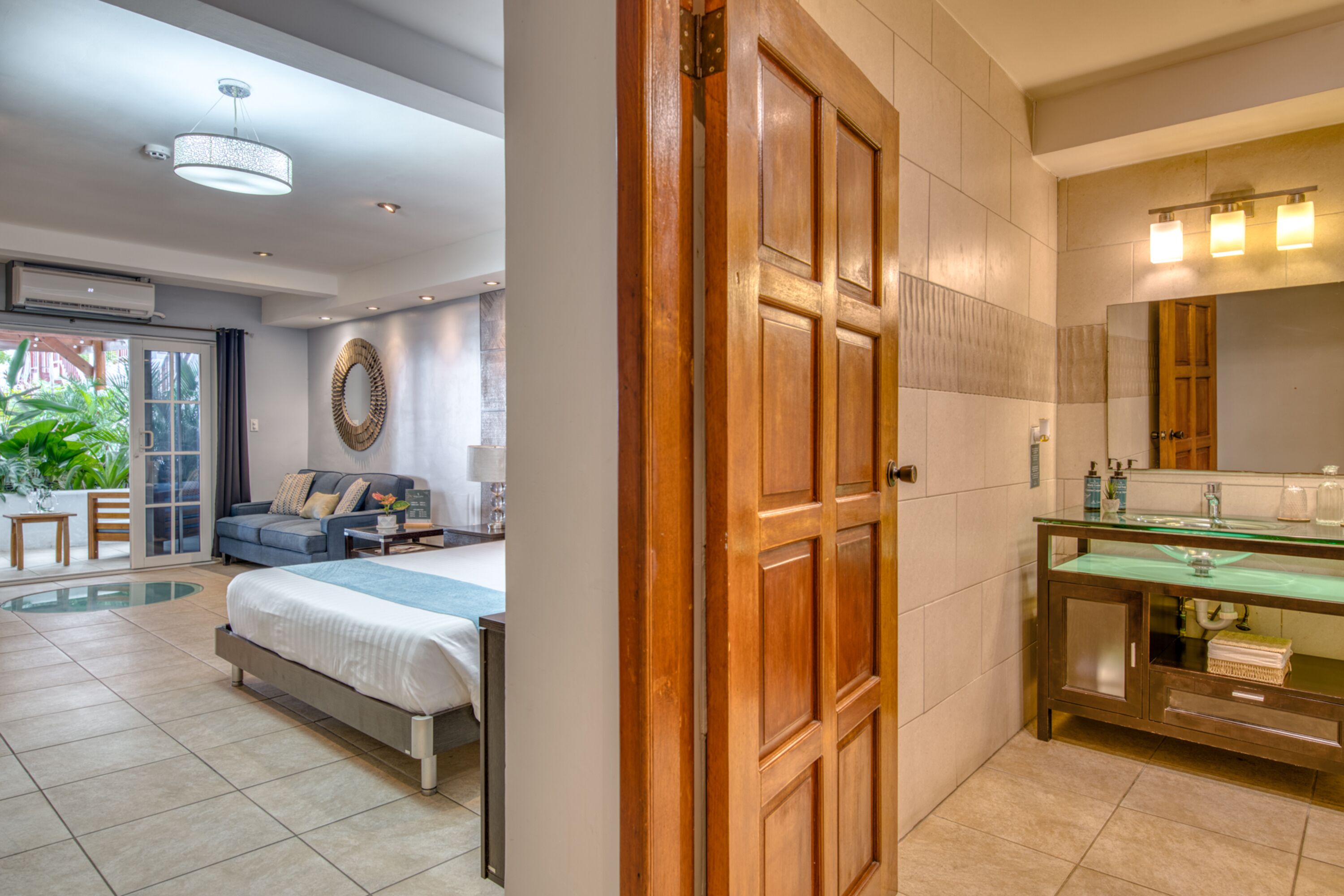 A hotel room with a bed, a sofa, a dining table, a large mirror, and a bathroom with a glass shower enclosure.