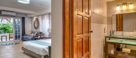 A hotel room with a bed, a sofa, a dining table, a large mirror, and a bathroom with a glass shower enclosure.