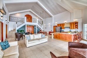 A spacious living area with a high ceiling, a staircase leading to an upper level, a kitchen with wooden cabinets, and a bar area. - Tropical Suites Hotel (Bocas del Toro)