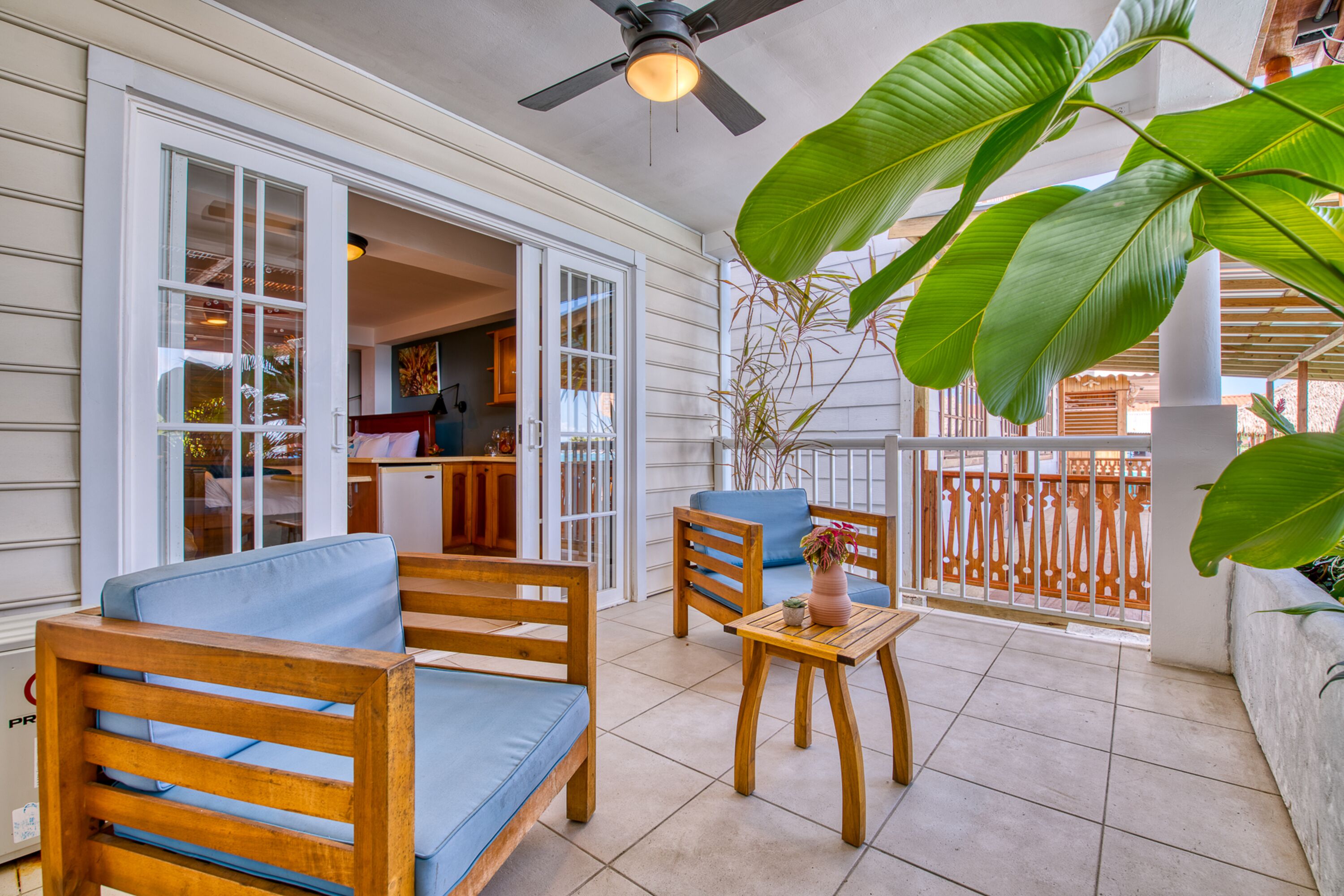A sunlit patio with wooden furniture, a ceiling fan, and large green plants.