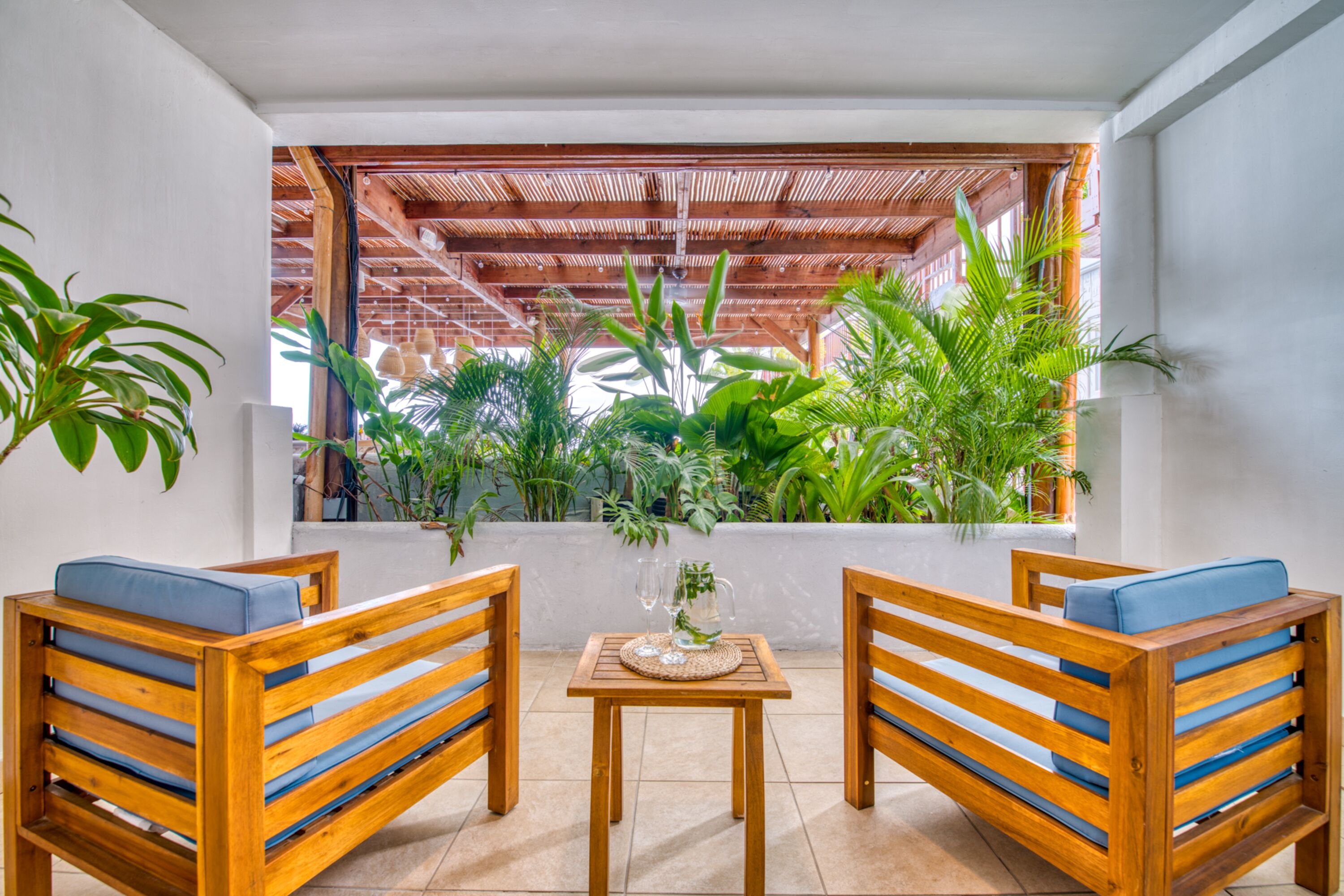 Un banco de madera con cojines azules, una mesita con una jarrón y vasos, y una vista de un jardín exuberante a través de un pérgola de madera.
