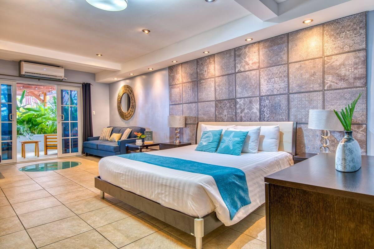 A modern hotel room with a large bed, a seating area with a sofa and chairs, a small table, and a decorative mirror.