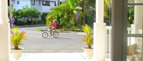 A covered entrance with a tiled floor, potted plants, and a view of a street with a person riding a bicycle.