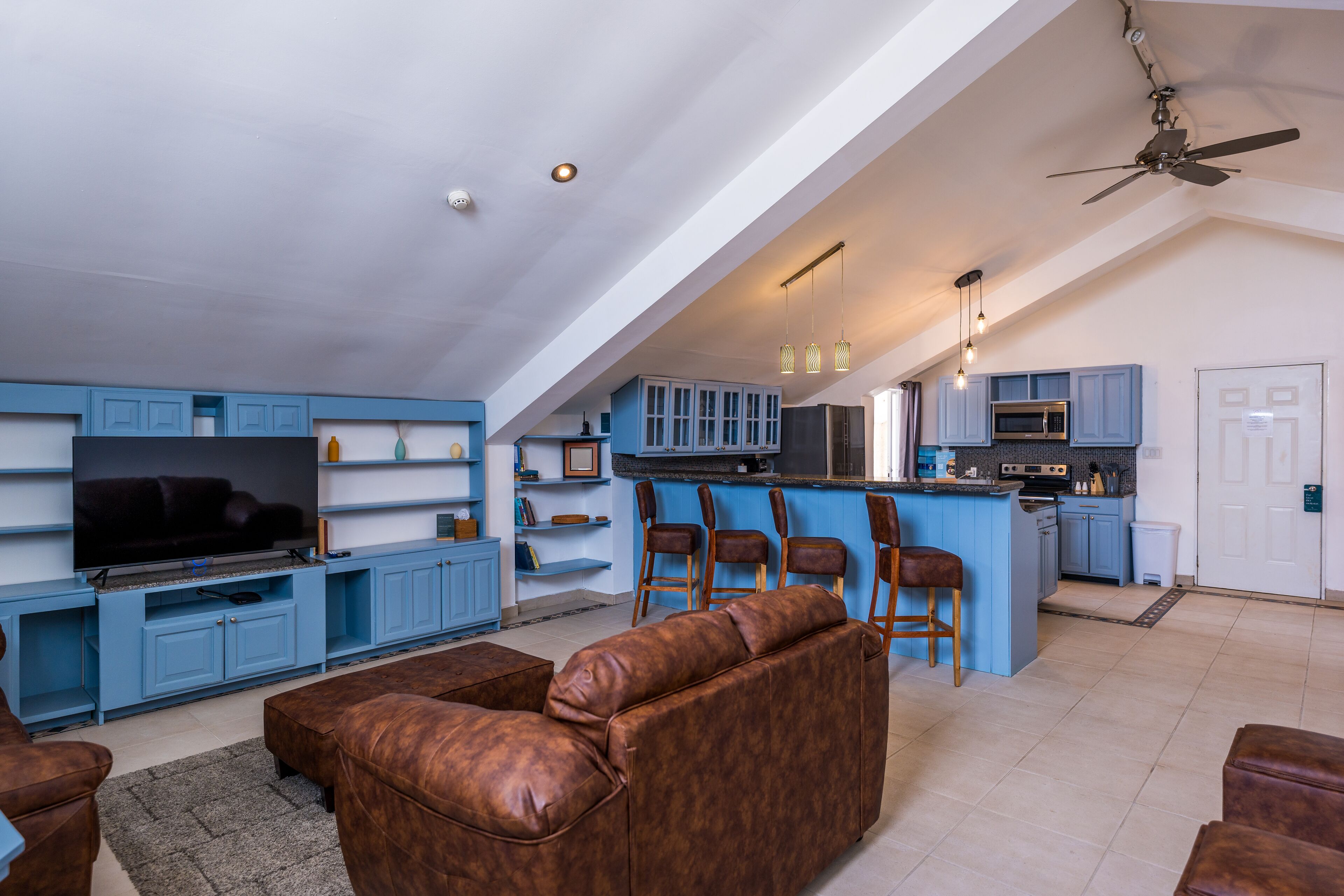 A spacious living area with a large flat-screen TV, a brown leather sofa, and a kitchen with blue cabinets and a bar area.