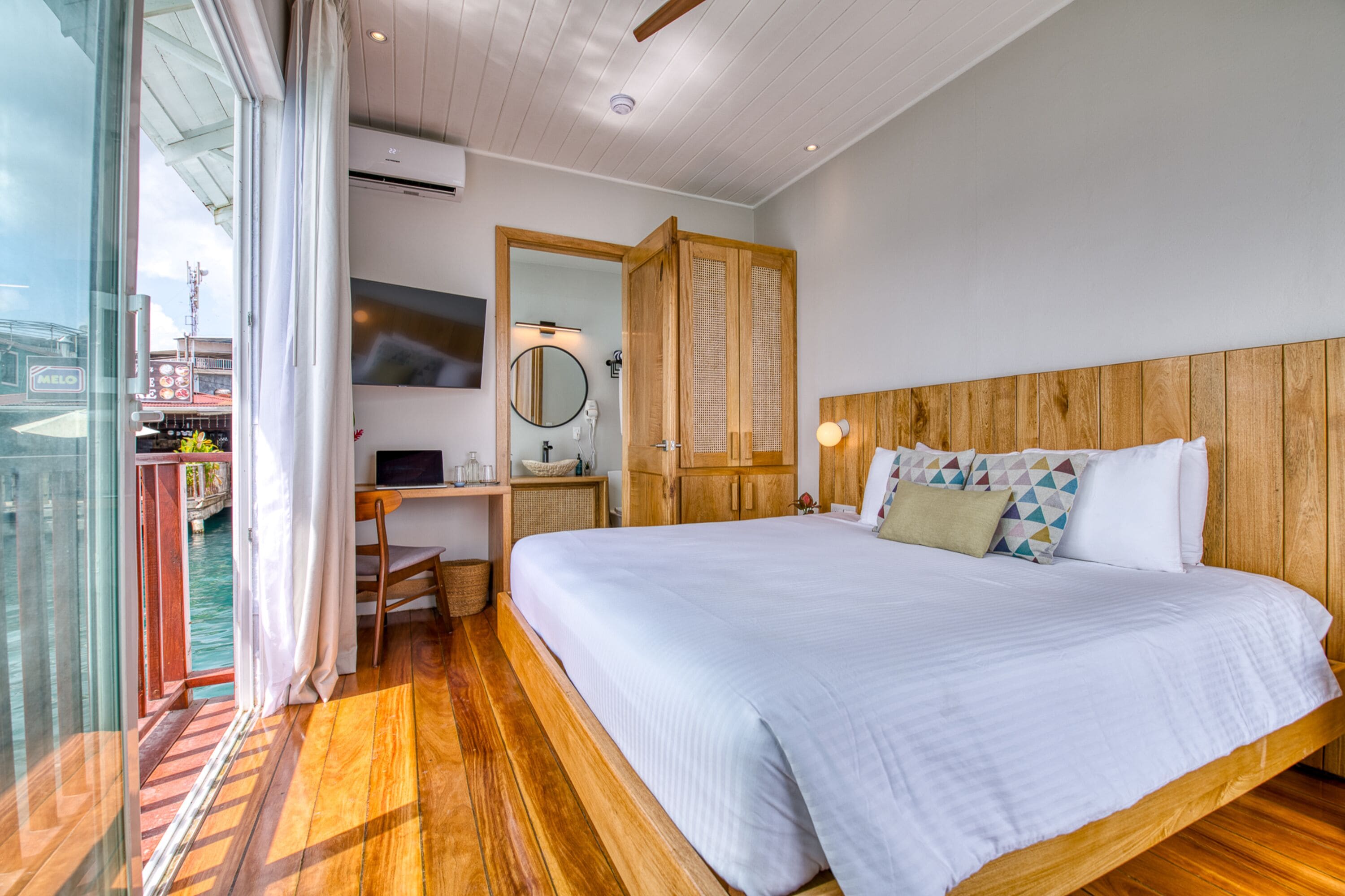 A bedroom with a wooden headboard, a large bed with white bedding, a desk with a computer, a mirror, and a view of a body of water through a glass door.