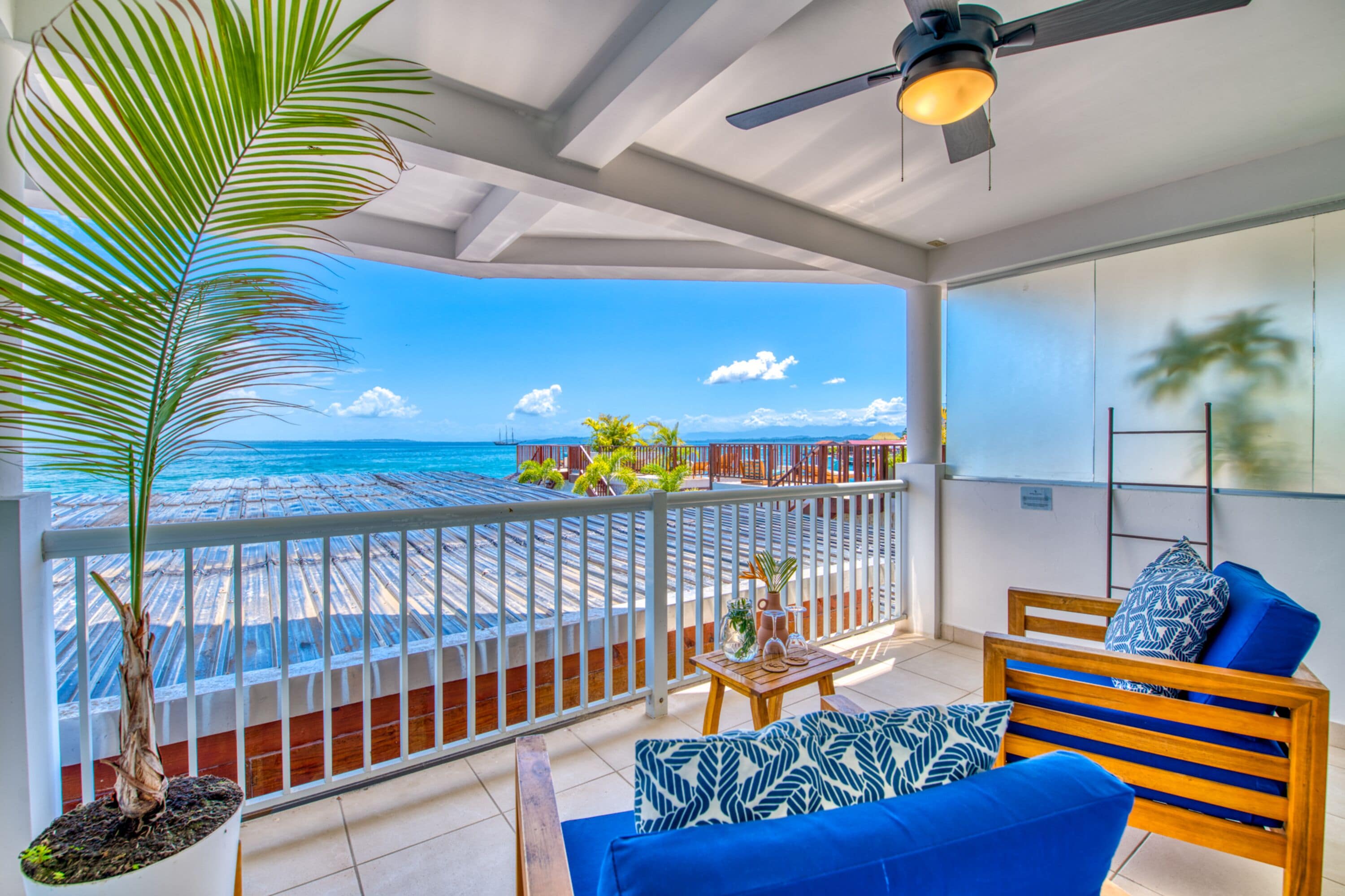 Un balcon avec vue sur la plage, un palmier et un coin salon avec une table et des coussins bleus.
