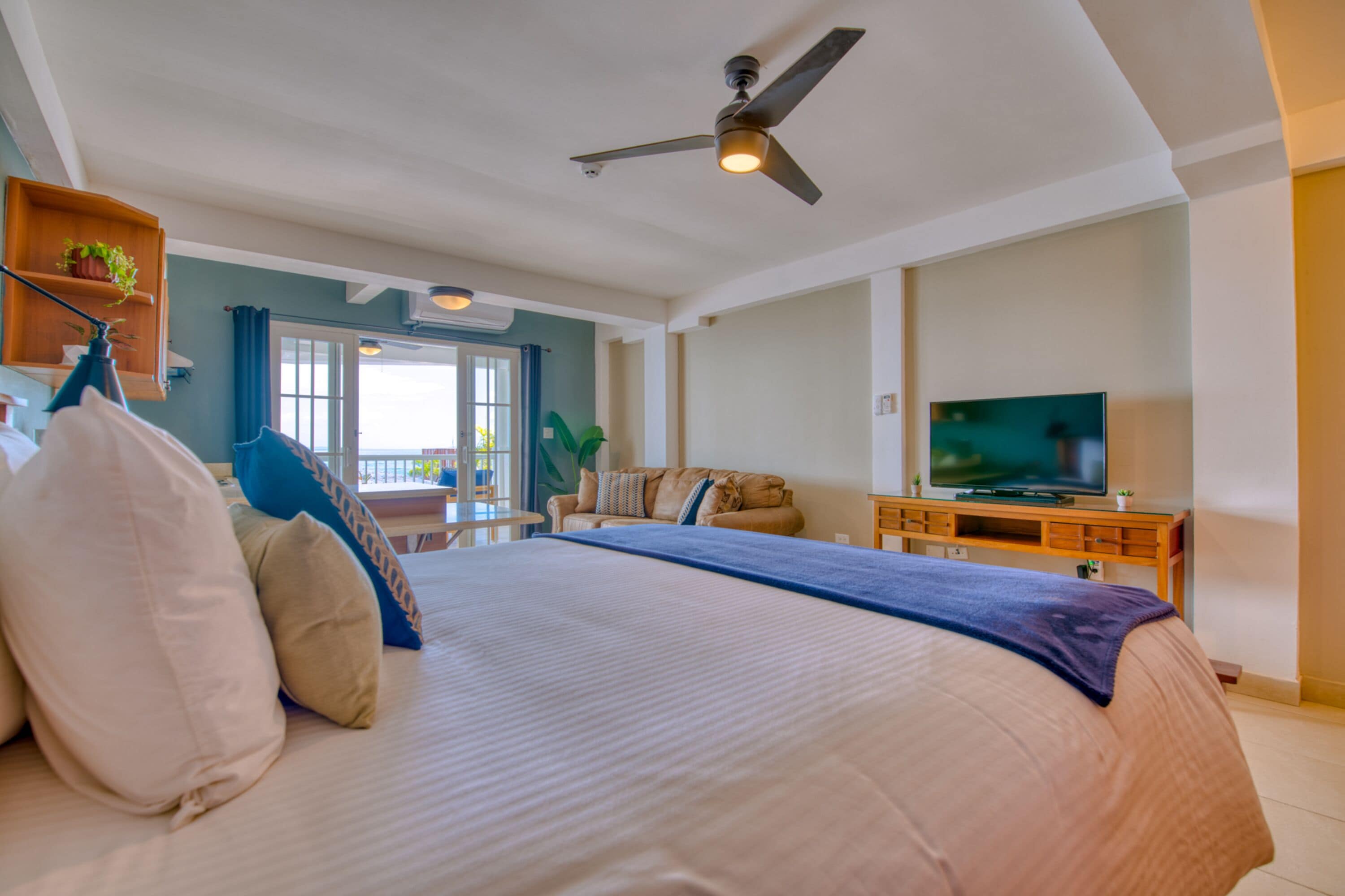 A spacious bedroom with a large bed, a ceiling fan, a television, and a balcony with a view.