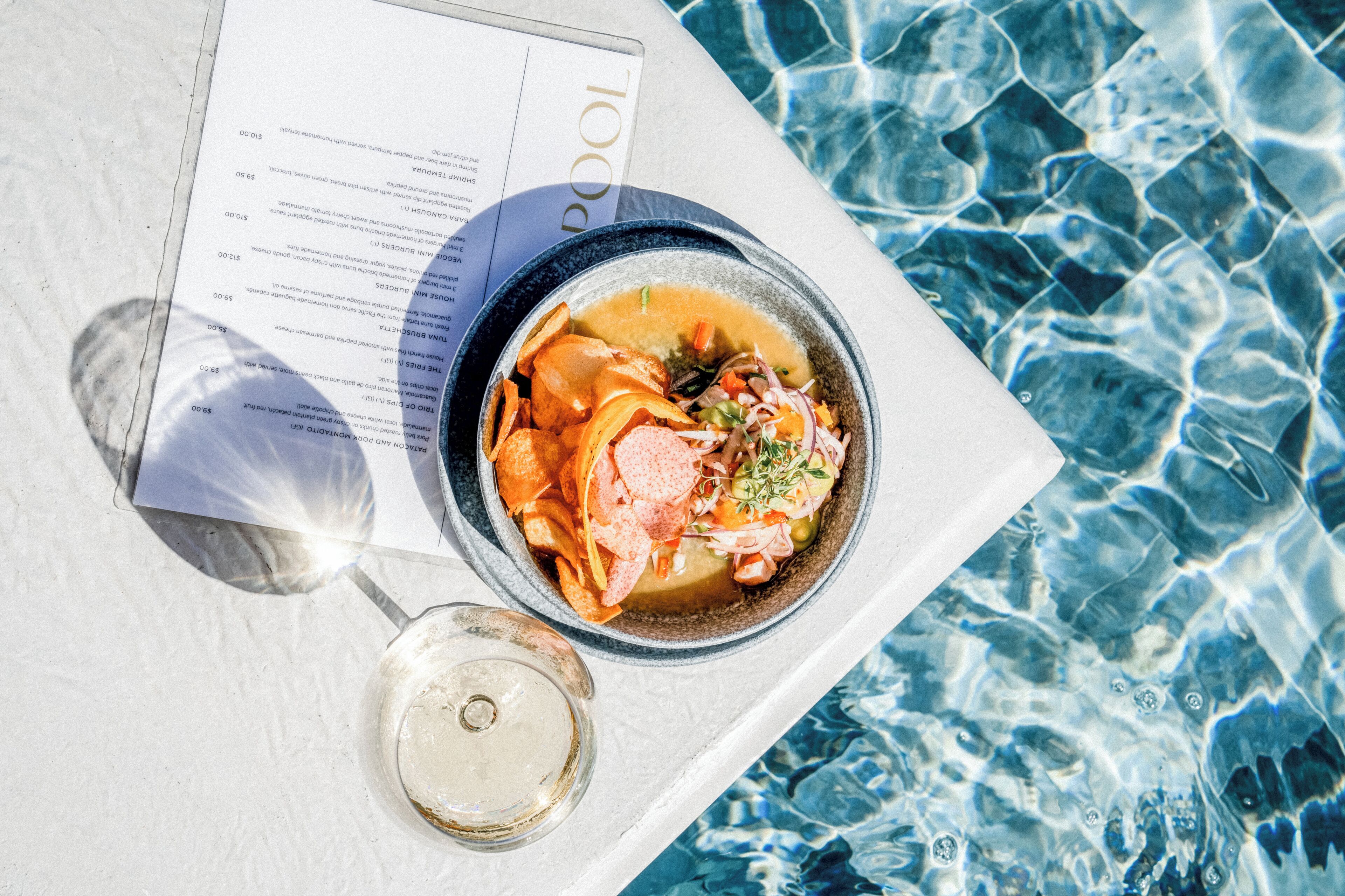 A dining table with a bowl of soup and chips, a menu, and a glass of water by a pool.
