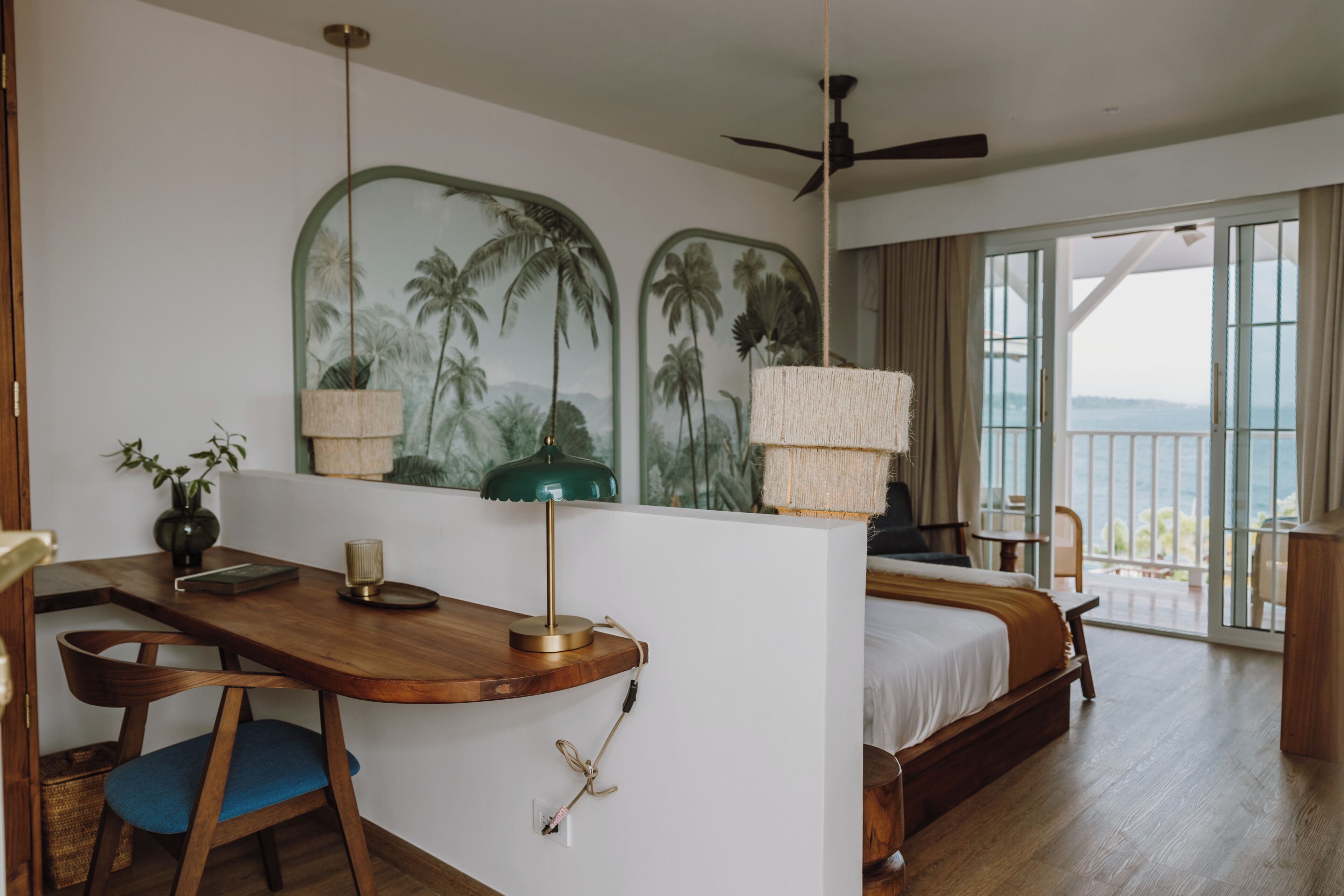 Habitación de hotel con escritorio de madera, cama, balcón con vistas y ventilador de techo.