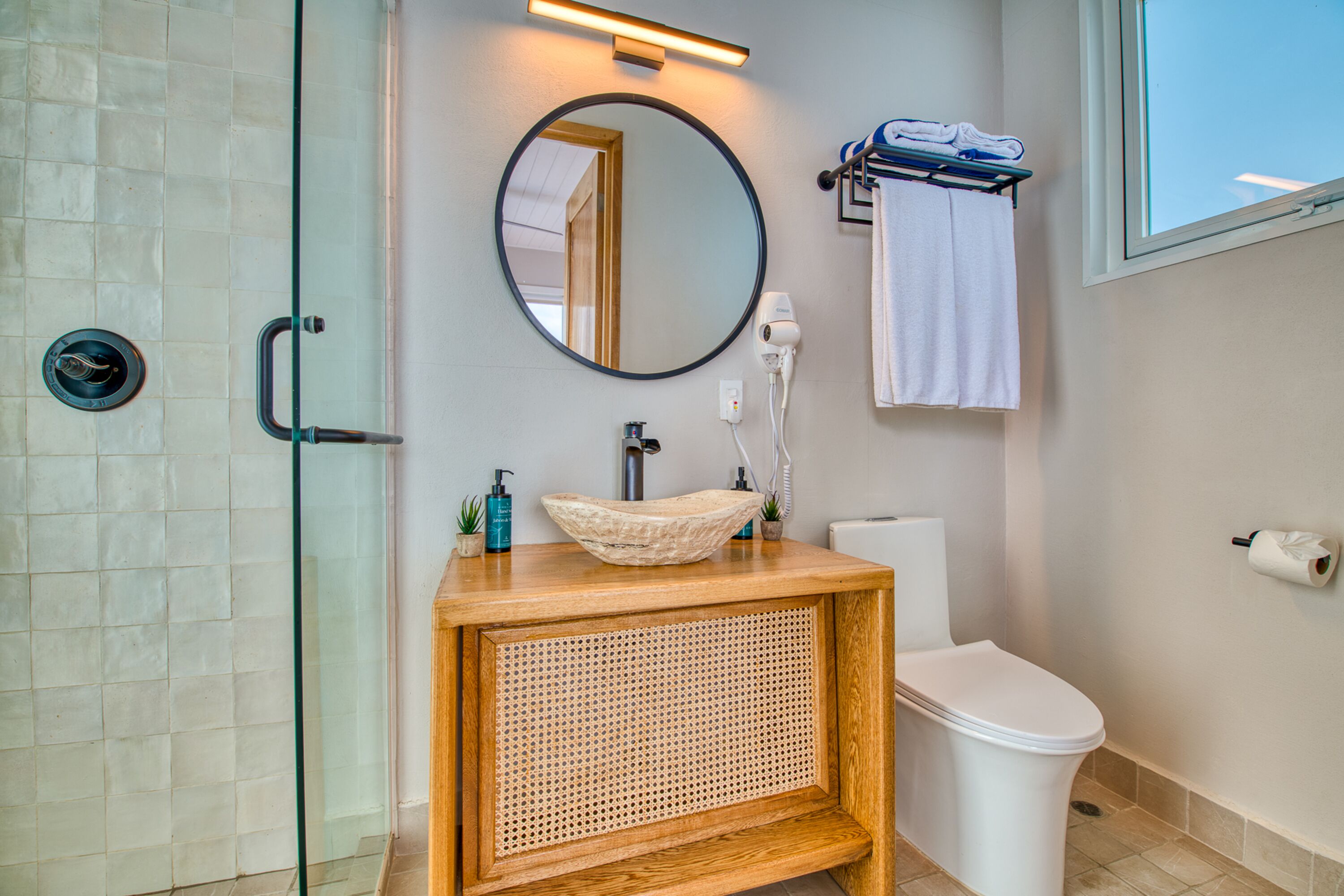 A bathroom with a glass shower enclosure, a wooden vanity with a woven front panel, a round mirror, a white toilet, and a towel rack with towels.