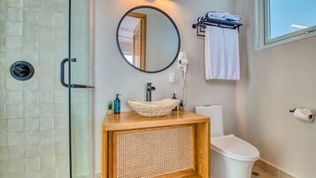 Une salle de bain avec une douche à parois de verre, un meuble-lavabo en bois avec un panneau avant tressé, un miroir rond, des toilettes blanches et un porte-serviettes avec des serviettes.