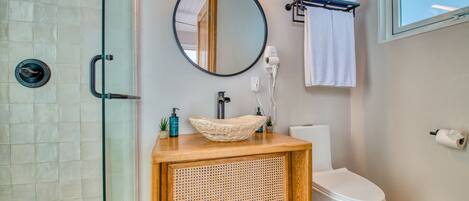 A bathroom with a glass shower enclosure, a wooden vanity with a woven front panel, a round mirror, a white toilet, and a towel rack with towels.