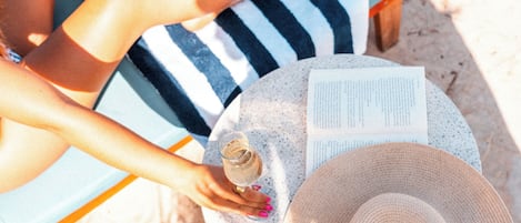 A person sitting on a striped beach chair with a book and a glass of drink on a round table.