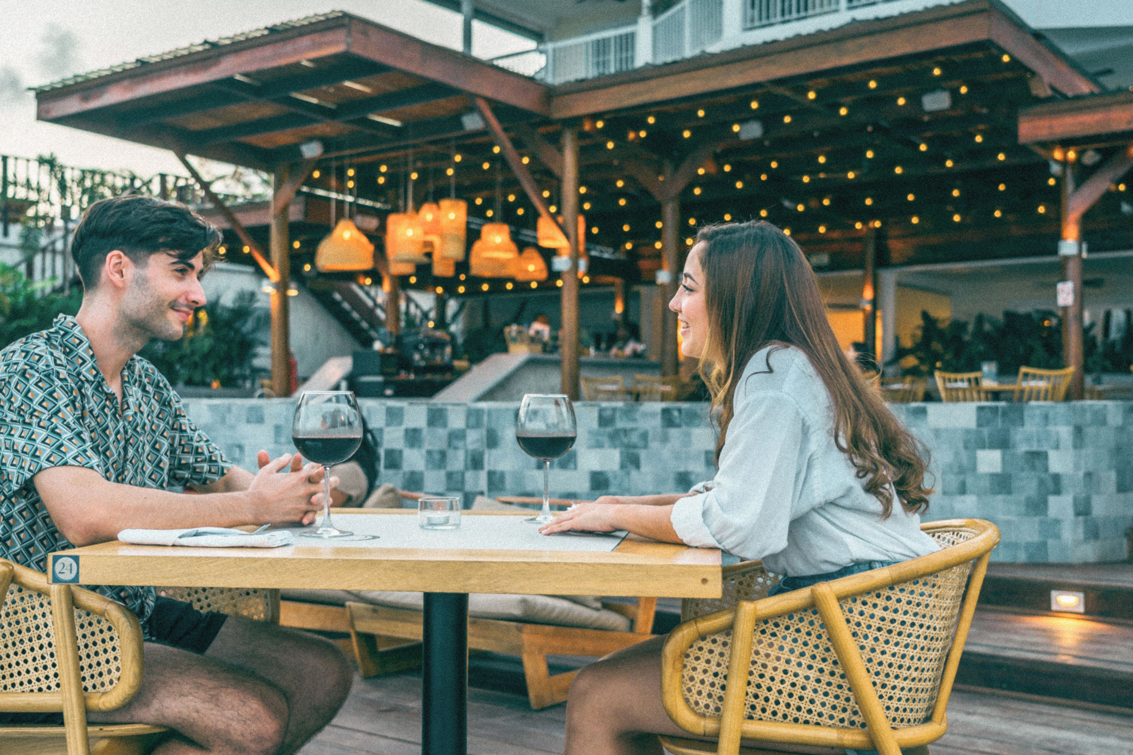 A couple dining at an outdoor restaurant with a checkered floor, wooden chairs, and hanging lights.