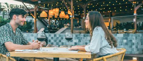 A couple dining at an outdoor restaurant with a checkered floor, wooden chairs, and hanging lights.