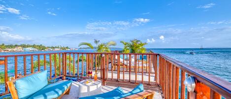 A wooden deck with two lounge chairs and a small table, overlooking a body of water with a distant ship and palm trees.