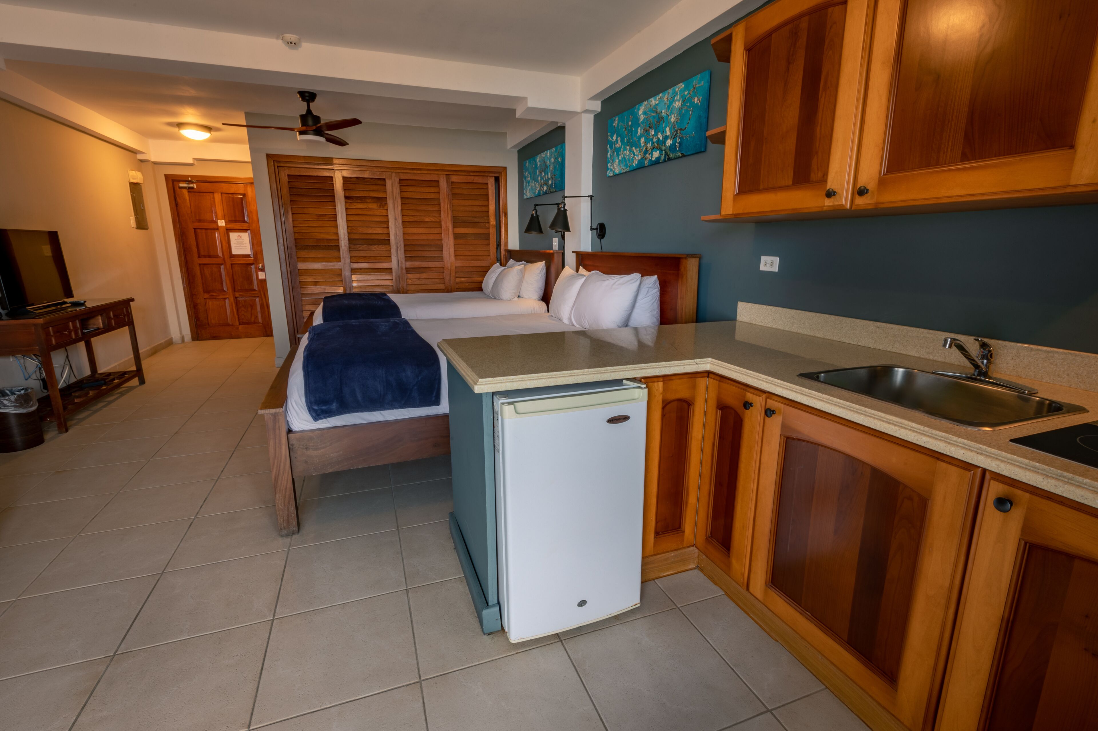 A compact hotel room with a bed, a small kitchenette, a ceiling fan, and wooden cabinets.