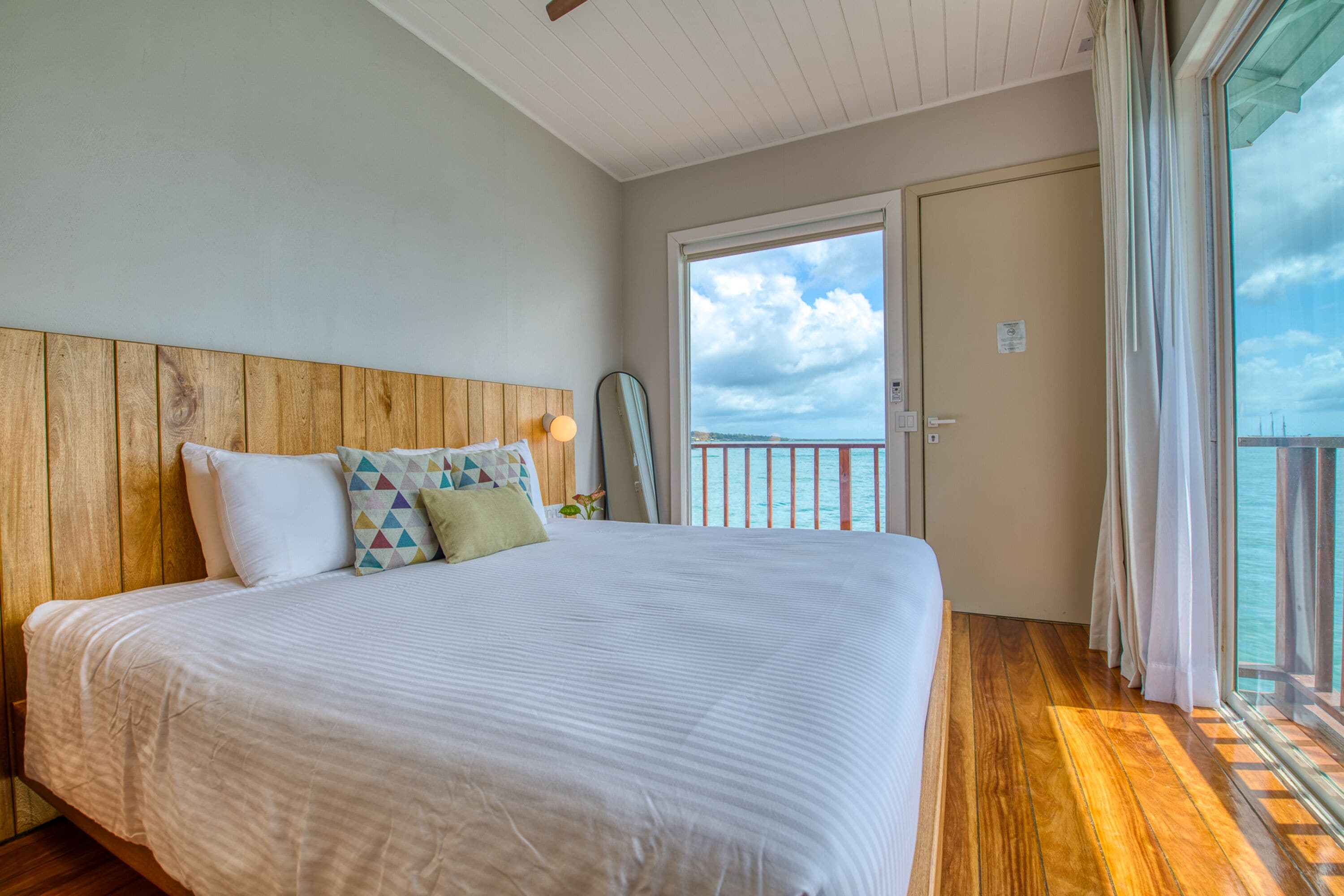 A bedroom with a wooden headboard, a large bed with white bedding, and a view of the sea through a sliding glass door.