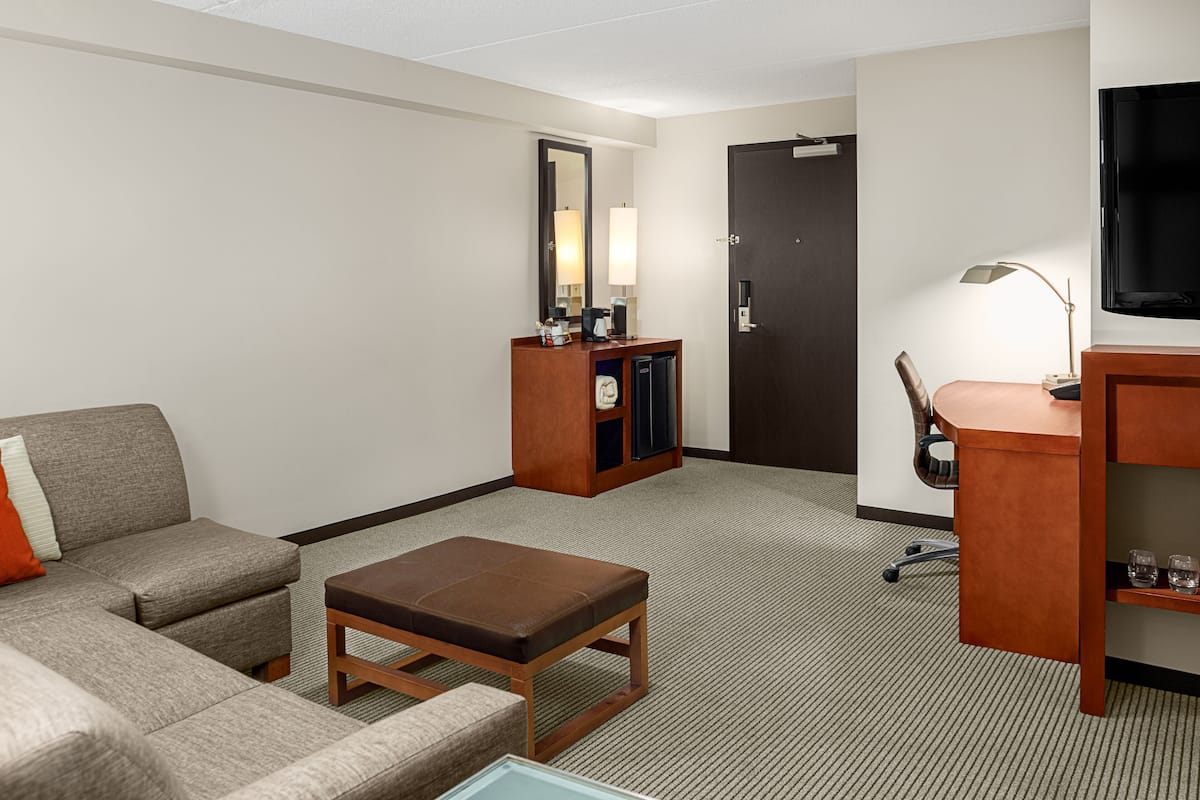 A hotel room with a sofa, a small table, a desk with a chair, a TV, a mirror, and a door.