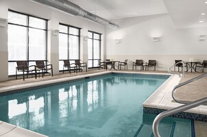 An indoor swimming pool with clear blue water, surrounded by chairs and tables, large windows with blinds, and a tiled floor.