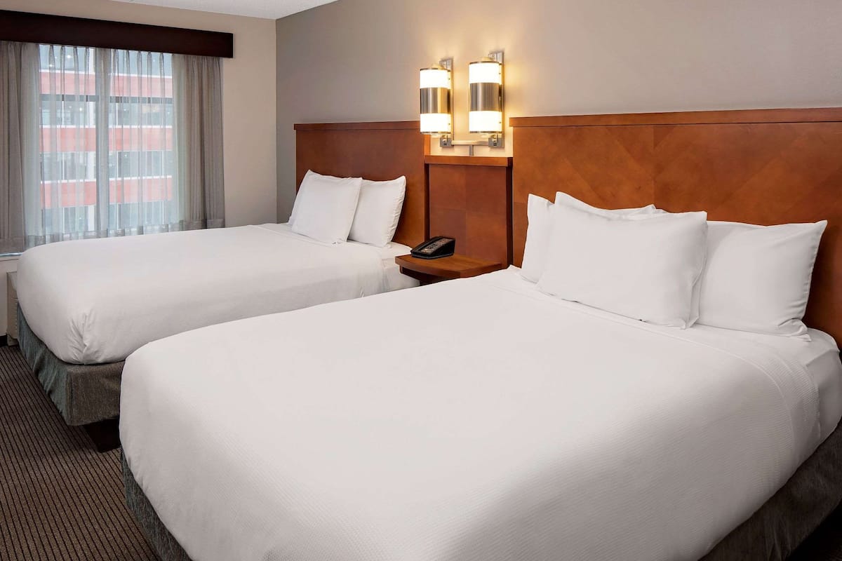 A hotel room with two beds, a wooden headboard, a bedside table with a phone, and a window with curtains.