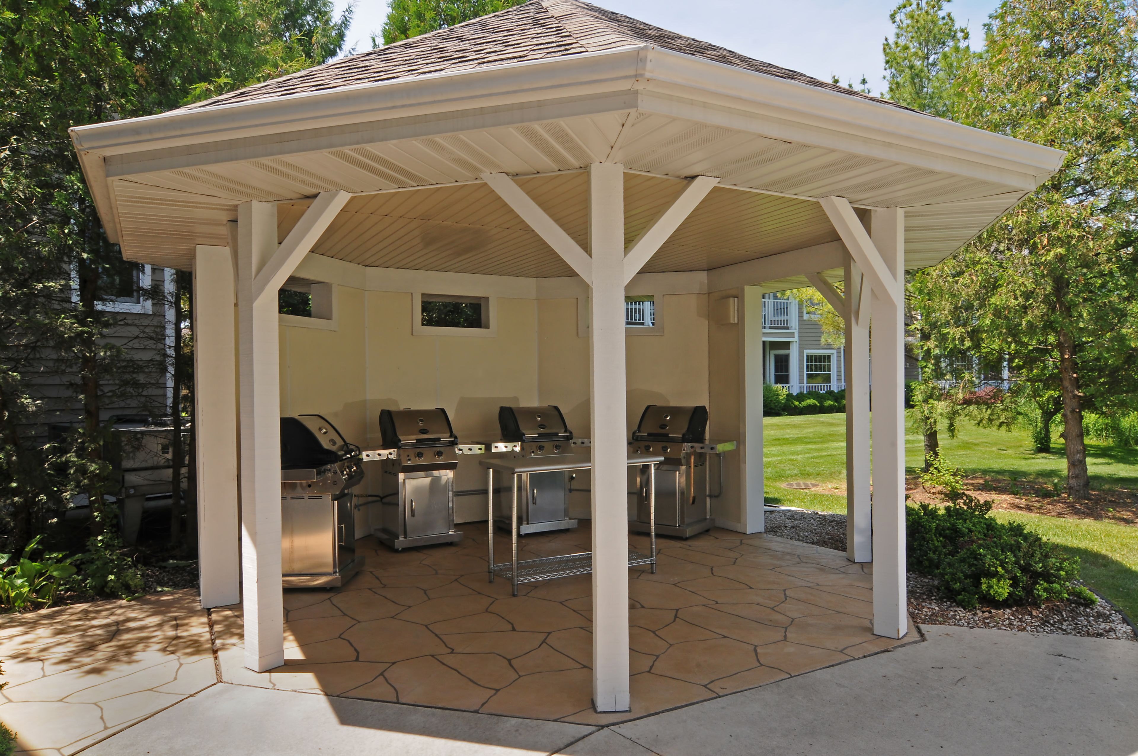 A covered outdoor grill area with three grills, a stone-tiled floor, and a white pergola structure.
