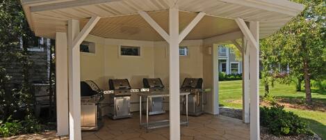 A covered outdoor grill area with three grills, a stone-tiled floor, and a white pergola structure.