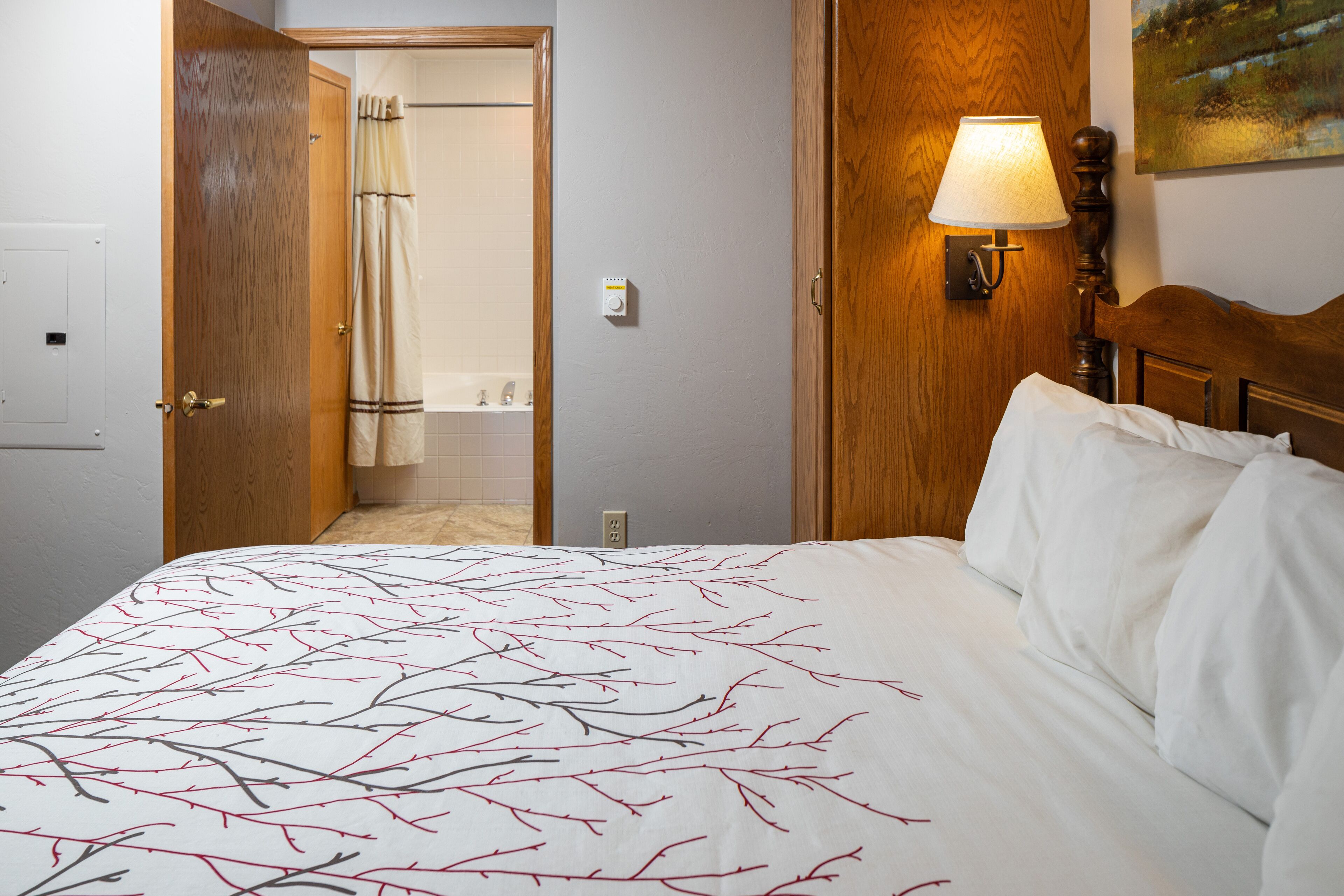 Ein Hotelzimmer mit einem Bett, einem hölzernen Kopfteil, einem Nachttisch mit Lampe und einem durch eine offene Tür einsehbaren Badezimmer.