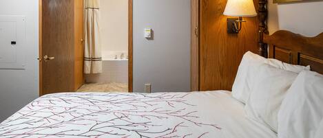 Ein Hotelzimmer mit einem Bett, einem hölzernen Kopfteil, einem Nachttisch mit Lampe und einem durch eine offene Tür einsehbaren Badezimmer.