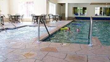 Piscina interior com chão de azulejos, posto de salva-vidas e mesa de bilhar com cadeiras.