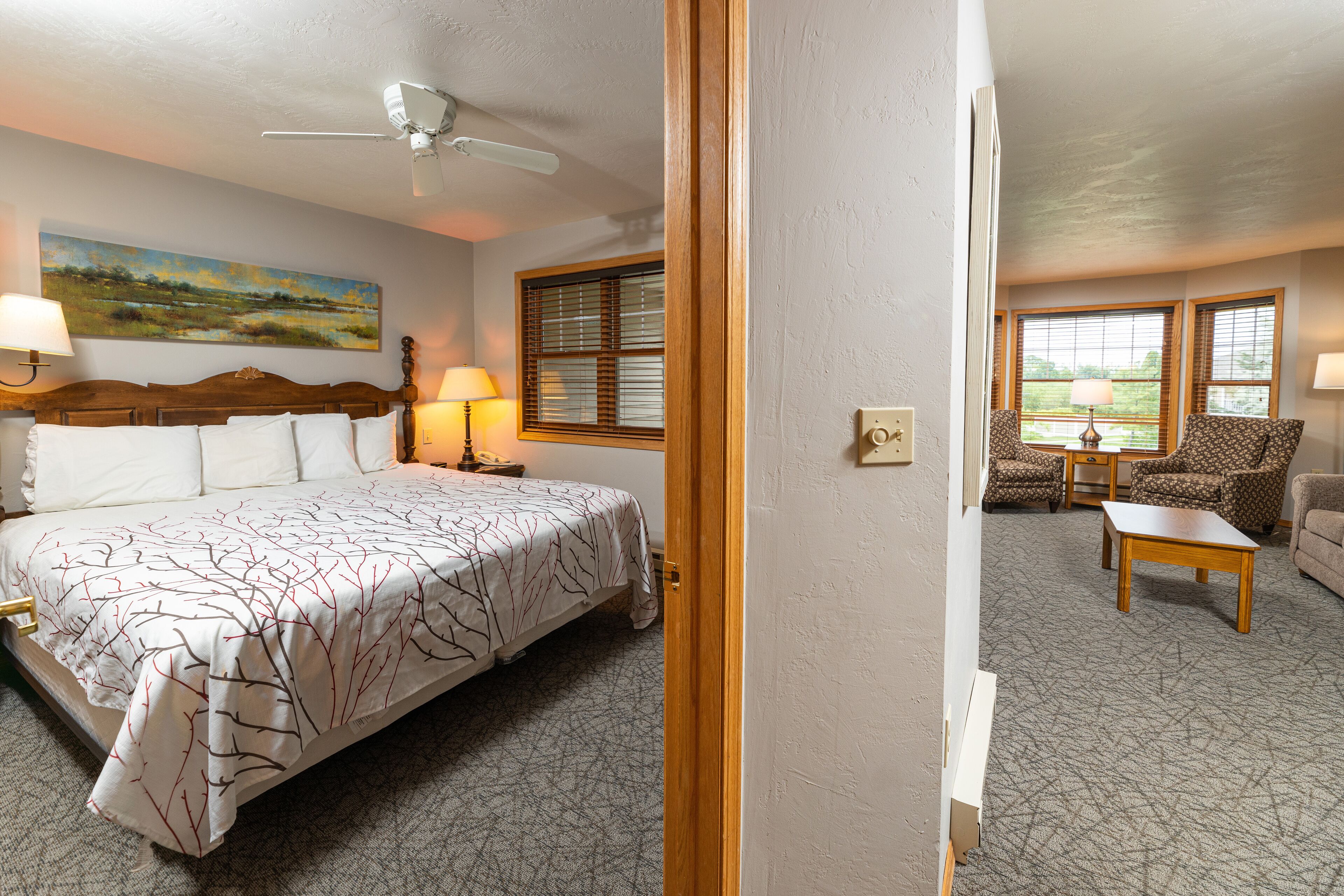 A bedroom with a bed, bedside lamps, a ceiling fan, a painting, and a view of the outdoors through a window.