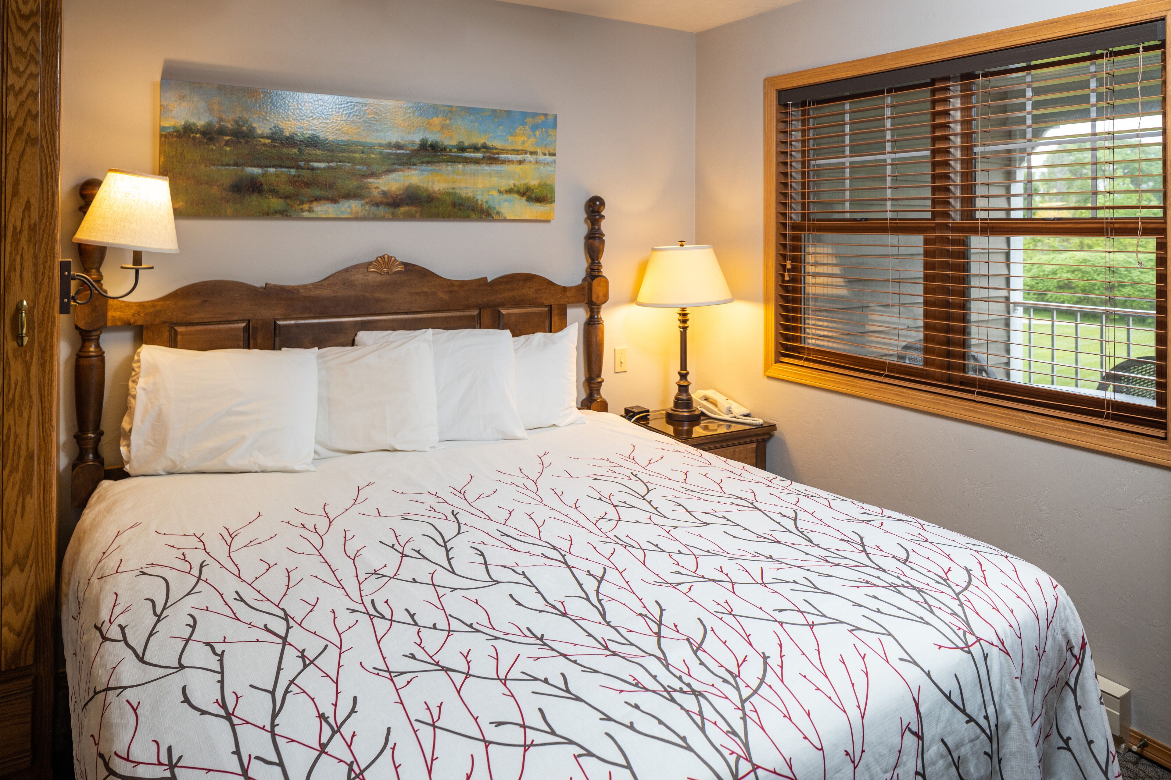 A neatly made bed with a white and red branch-patterned bedspread, a wooden headboard, two bedside lamps, and a painting of a landscape on the wall.