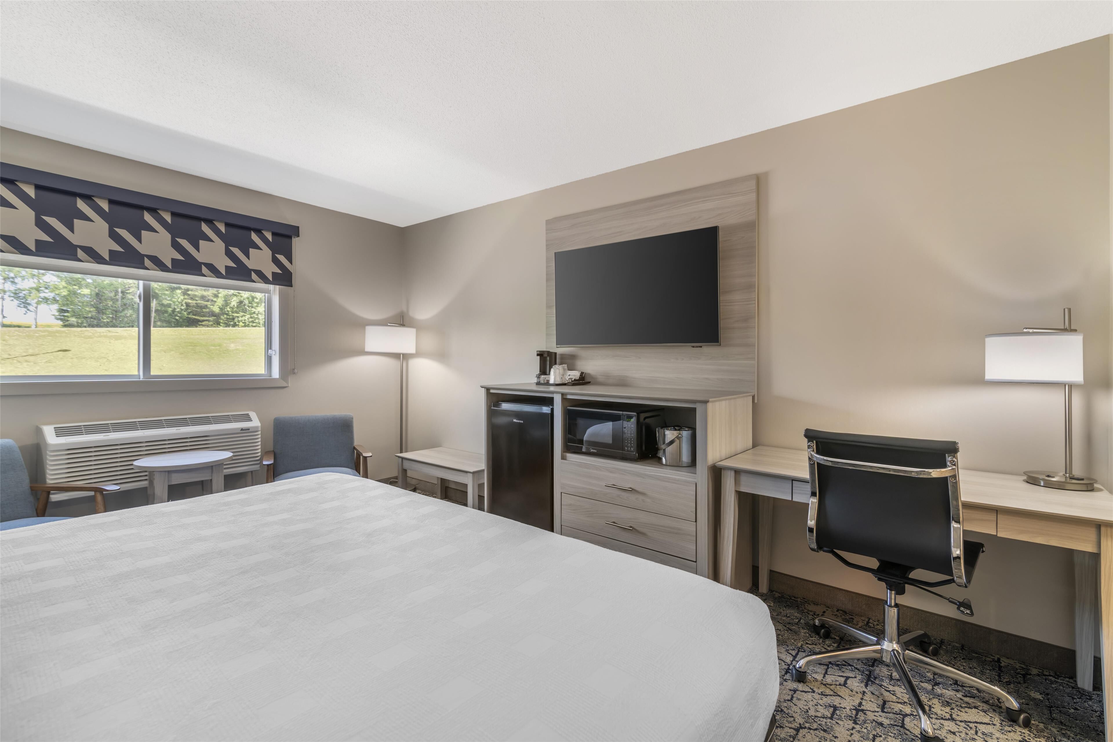 A hotel room with a large bed, a desk with a computer, a chair, a TV mounted on the wall, and a window with a view of the outdoors.